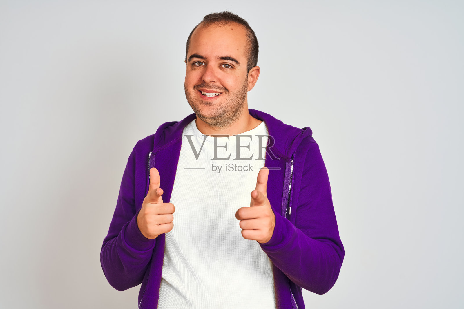 年轻男子穿着紫色的运动衫站在孤立的白色背景指向相机与快乐和有趣的脸。良好的能量和共鸣。照片摄影图片