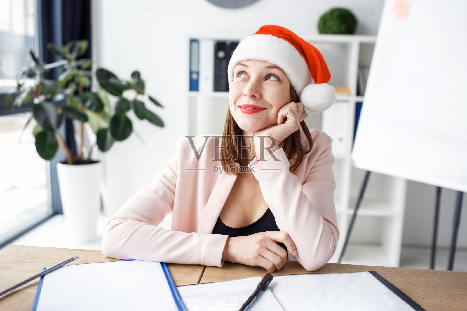 梦幻般积极而快乐的年轻女子抬头一笑。戴红帽子。节日的气氛中。庆祝新年或圣诞节。独自一人在工作。坐在办公室。照片摄影图片