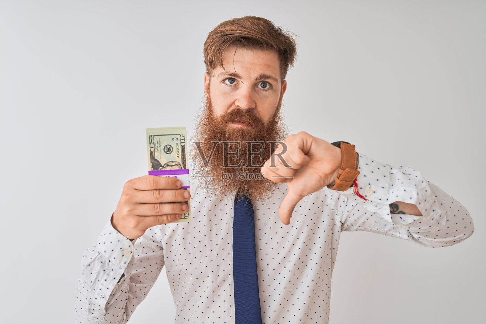 年轻的红发爱尔兰商人拿着美元站在孤立的白人背景上,面带愤怒,表示不喜欢,用拇指表示拒绝照片摄影图片