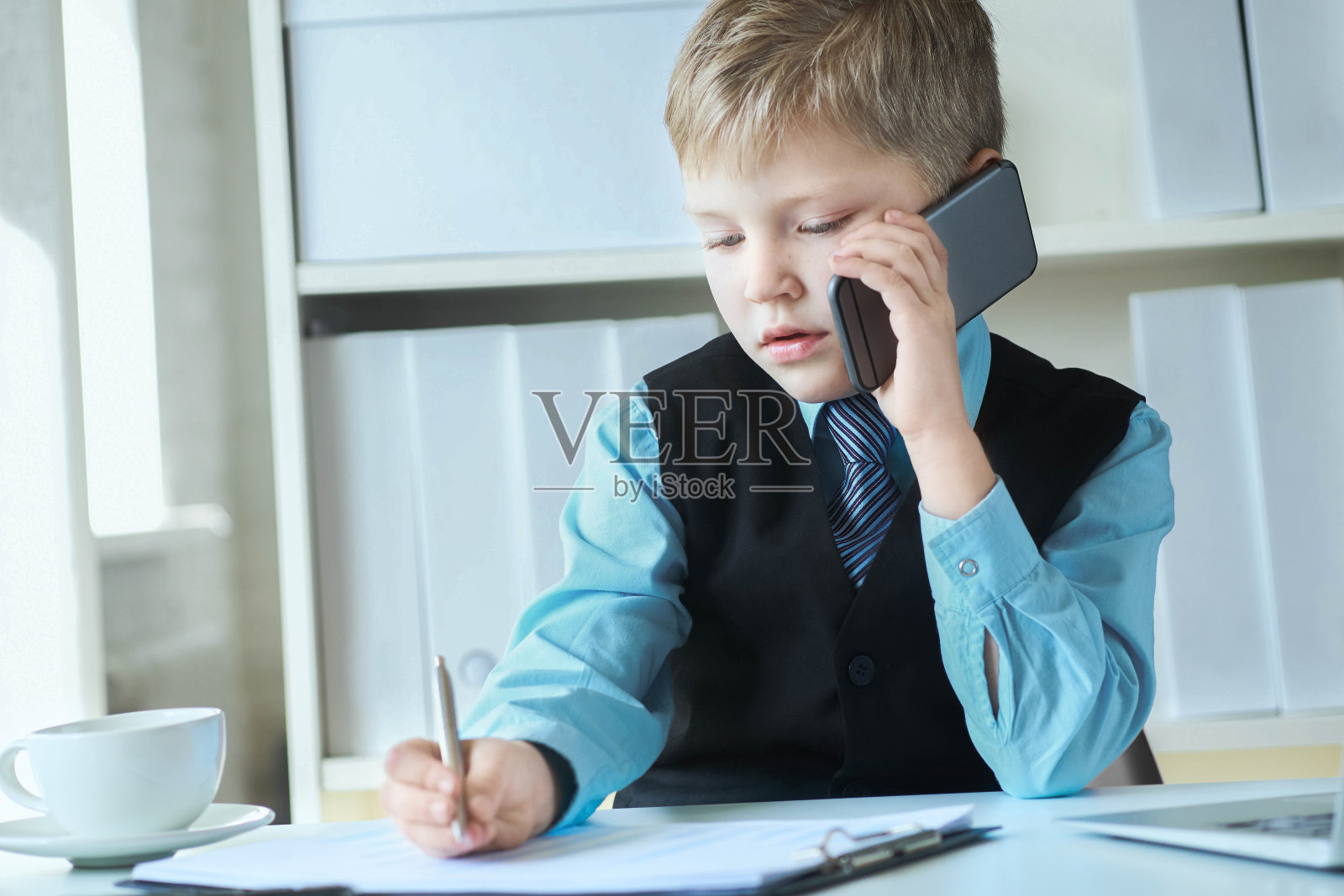 小自信的行政商人老板男孩做笔记和讲电话坐在桌子上用笔记本电脑在办公室。照片摄影图片