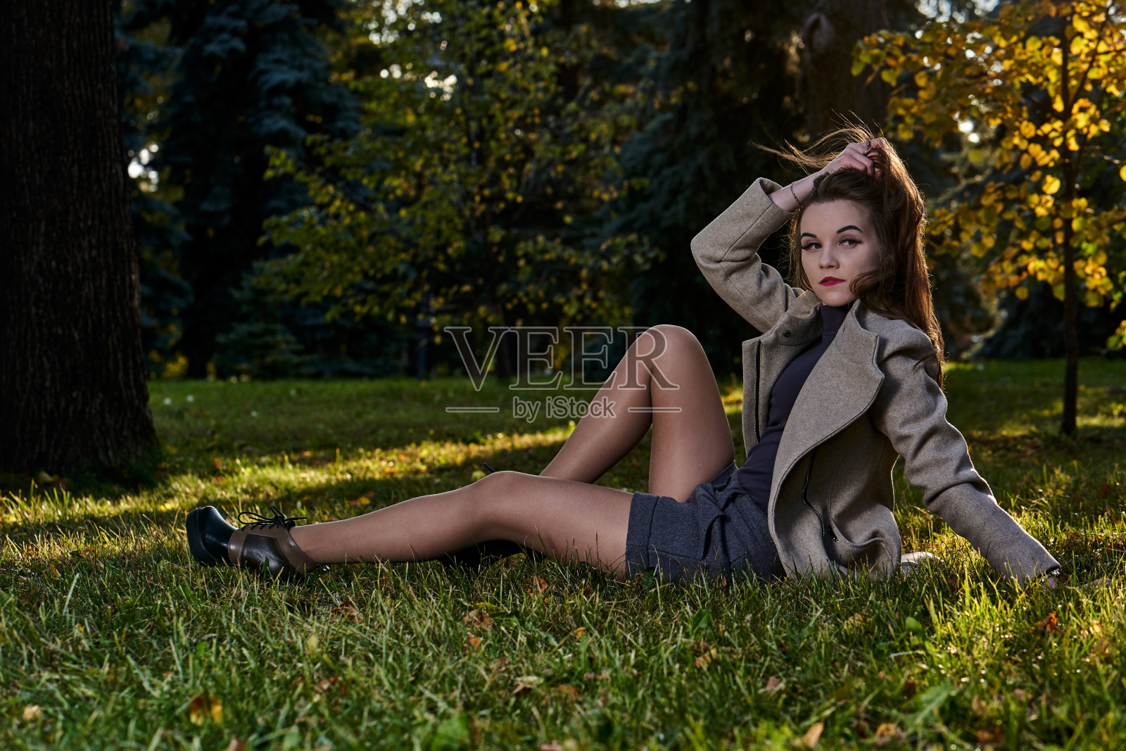 在一个阳光明媚的秋夜,一位穿着羊毛大衣的长发年轻女子在公园里散步。坐在草地上休息的女人。照片摄影图片