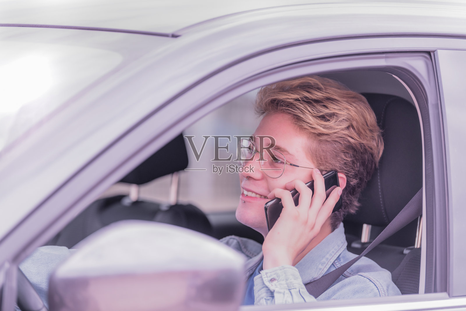 金发男孩坐在车里,眼睛直视前方,微笑着讲电话。照片摄影图片