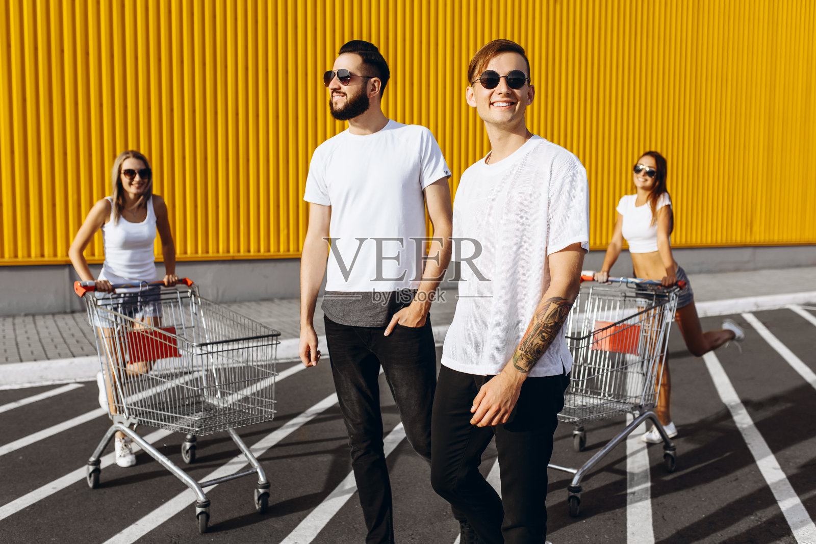 年轻人的公司,时髦的女孩和男孩,朋友有乐趣与购物车,户外超市附近在阳光明媚的天气照片摄影图片