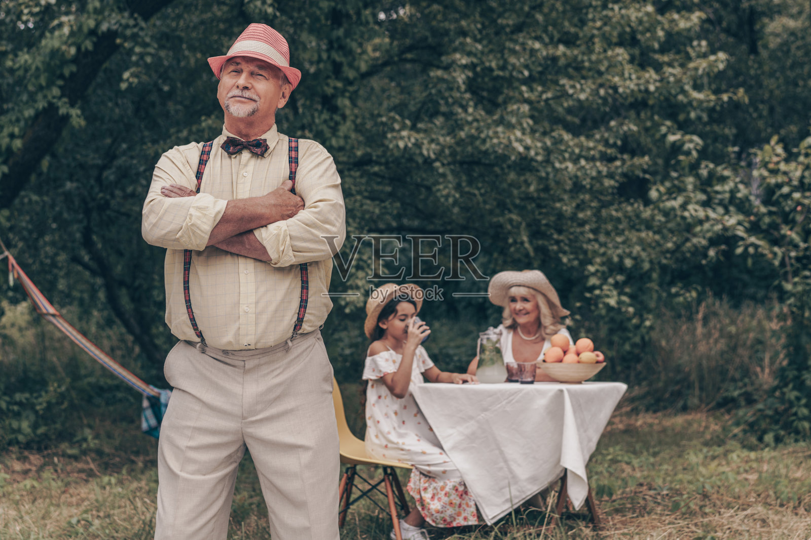 帅气的爷爷穿着红帽子、衬衫、裤子和蝴蝶结站在前台。后面坐在桌子上的老奶奶和年轻的孙女微笑着。幸福的全家福照片摄影图片