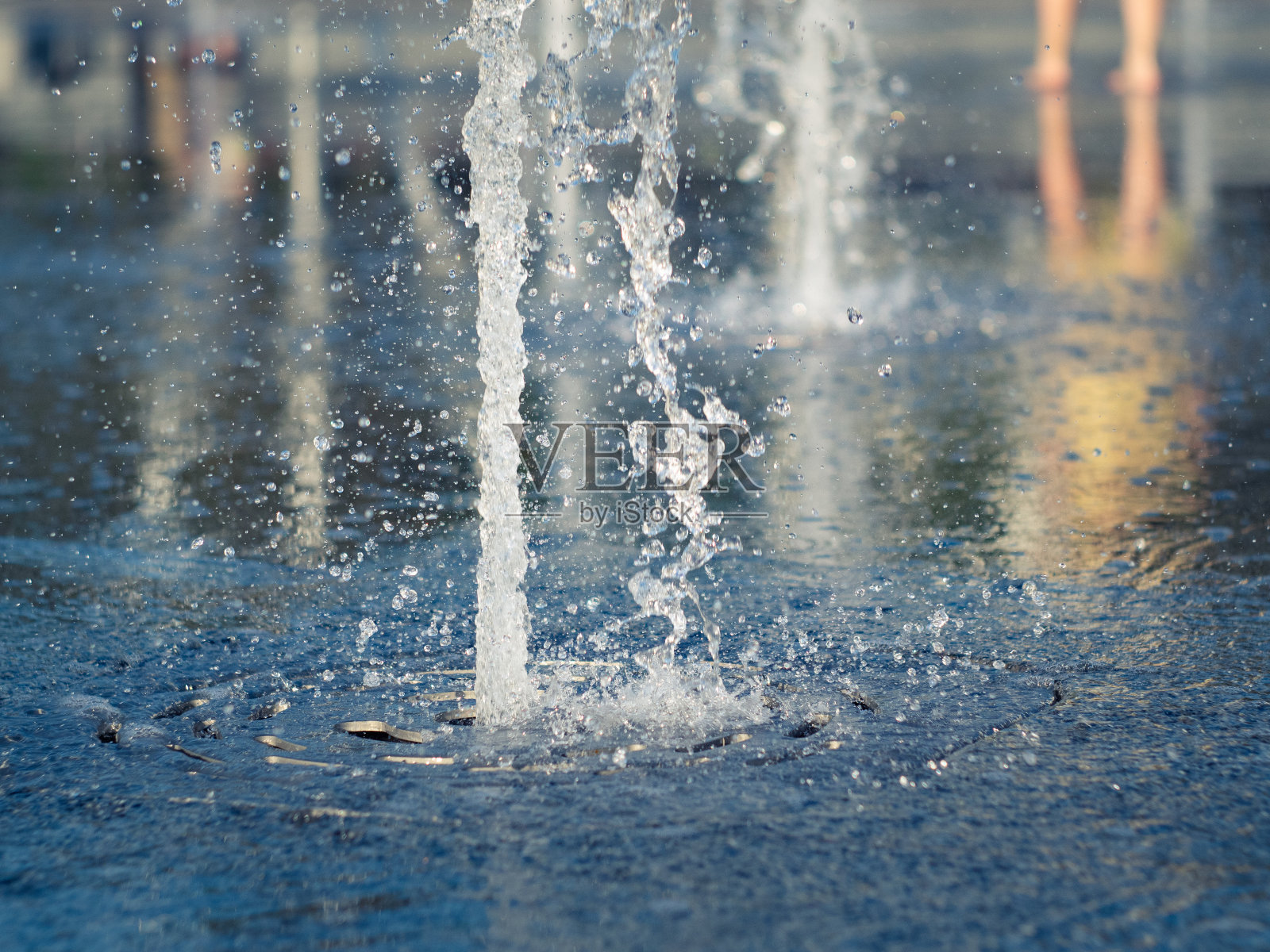 城市广场上的干喷泉,近景。水流溅在喷水池上,喷溅出许多水珠。孩子们在喷泉旁赤着脚。选择软焦点。模糊的背景。照片摄影图片