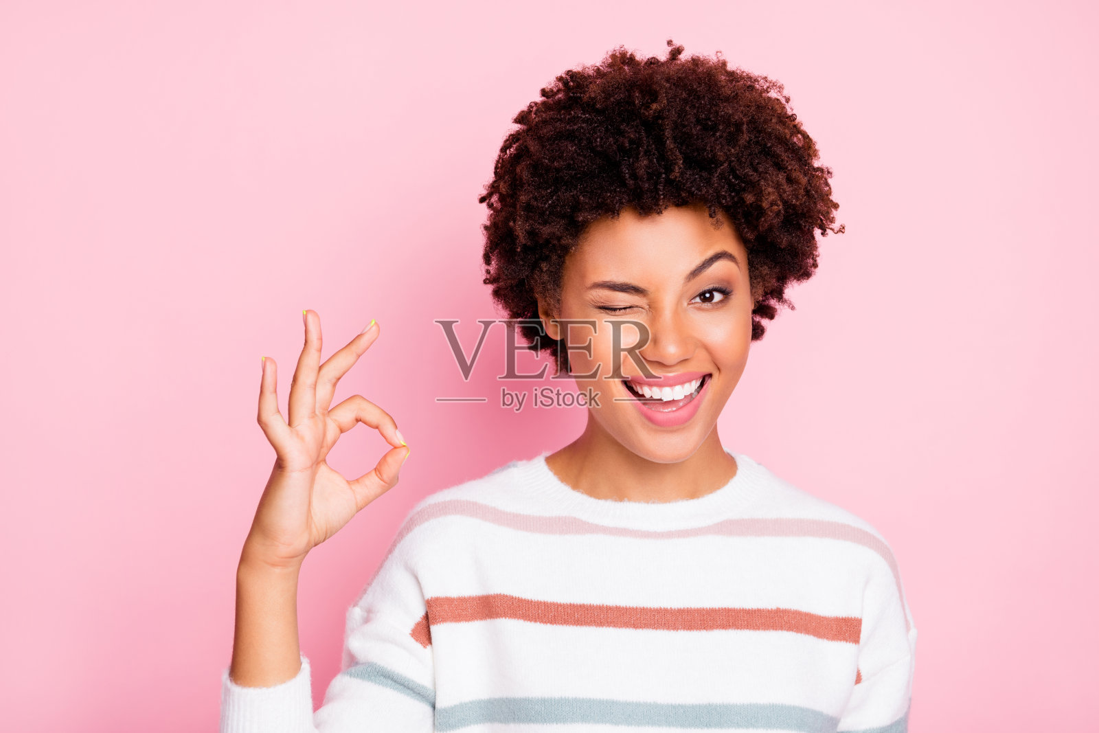 照片漂亮的黑皮肤女士眨眼睛显示ok符号完全同意购物价格穿白色条纹套头衫孤立的淡粉色背景照片摄影图片