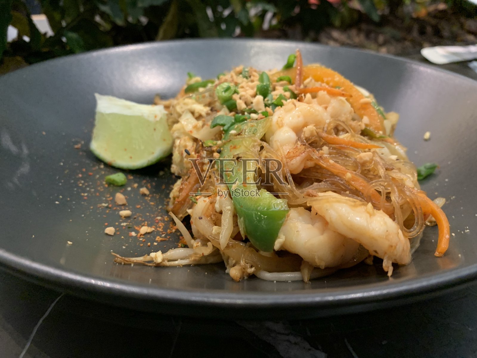 玻璃纸面条或玻璃粉与虾,花生,大蒜,泰国香料,甜椒和一片酸橙在一个黑色的圆盘子。背景用鲜花。照片摄影图片