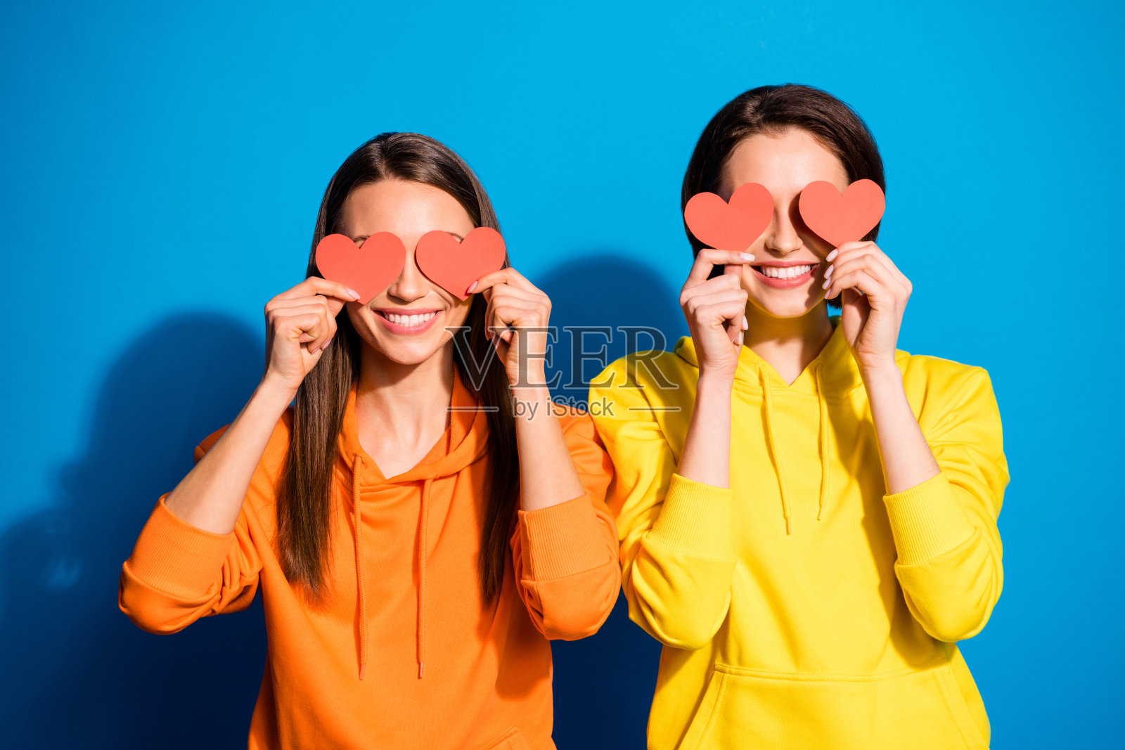 照片中的两个漂亮的两个女士拿着小红纸心隐藏眼睛表达积极的反应穿休闲的黄色连帽衫孤立的蓝色背景照片摄影图片