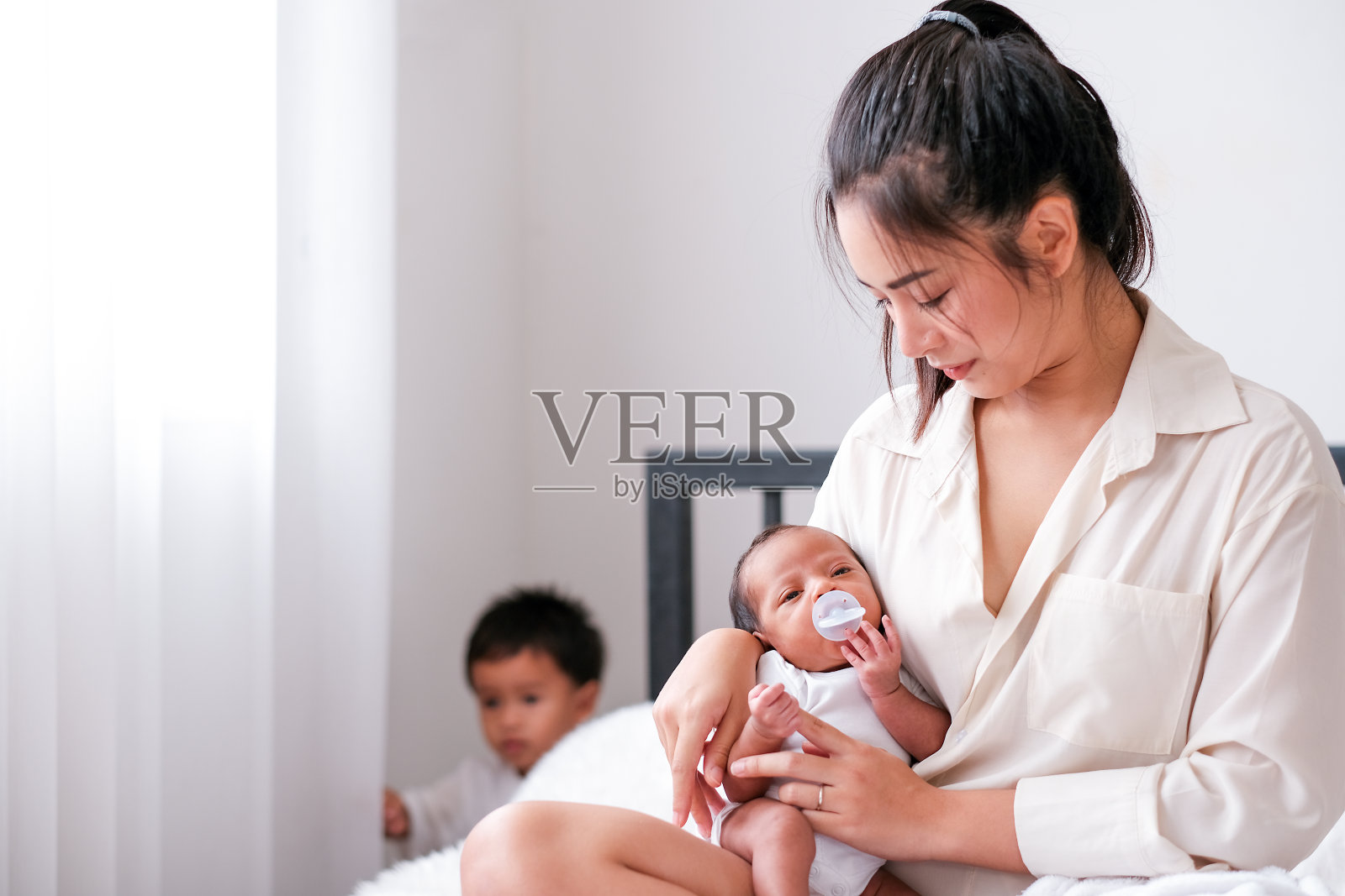 亚洲母亲抱着刚出生的婴儿,和儿子一起坐在卧室的床上照片摄影图片