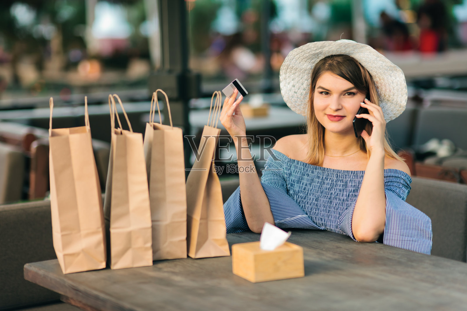 在线购物。一名兴高采烈的年轻女子坐在户外咖啡馆里,手里拿着银行卡,一边讲电话一边聊天照片摄影图片