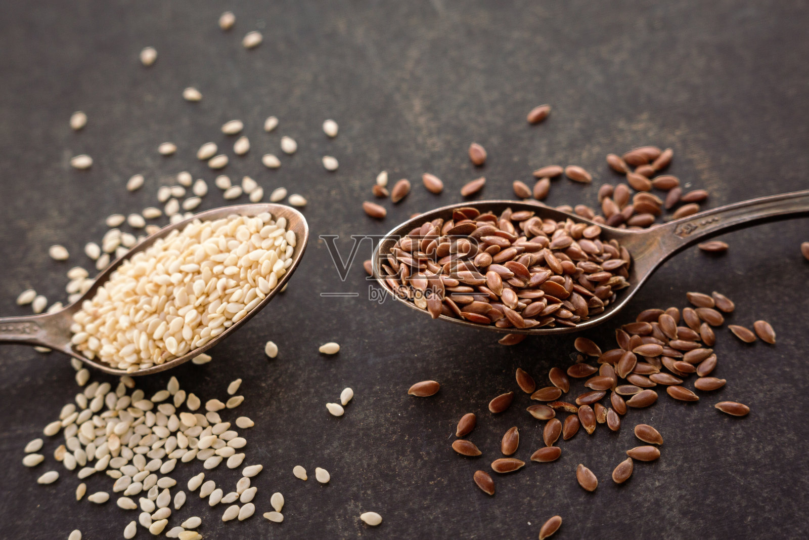 把芝麻和亚麻籽放在勺子里,放在深色的背景上。照片摄影图片