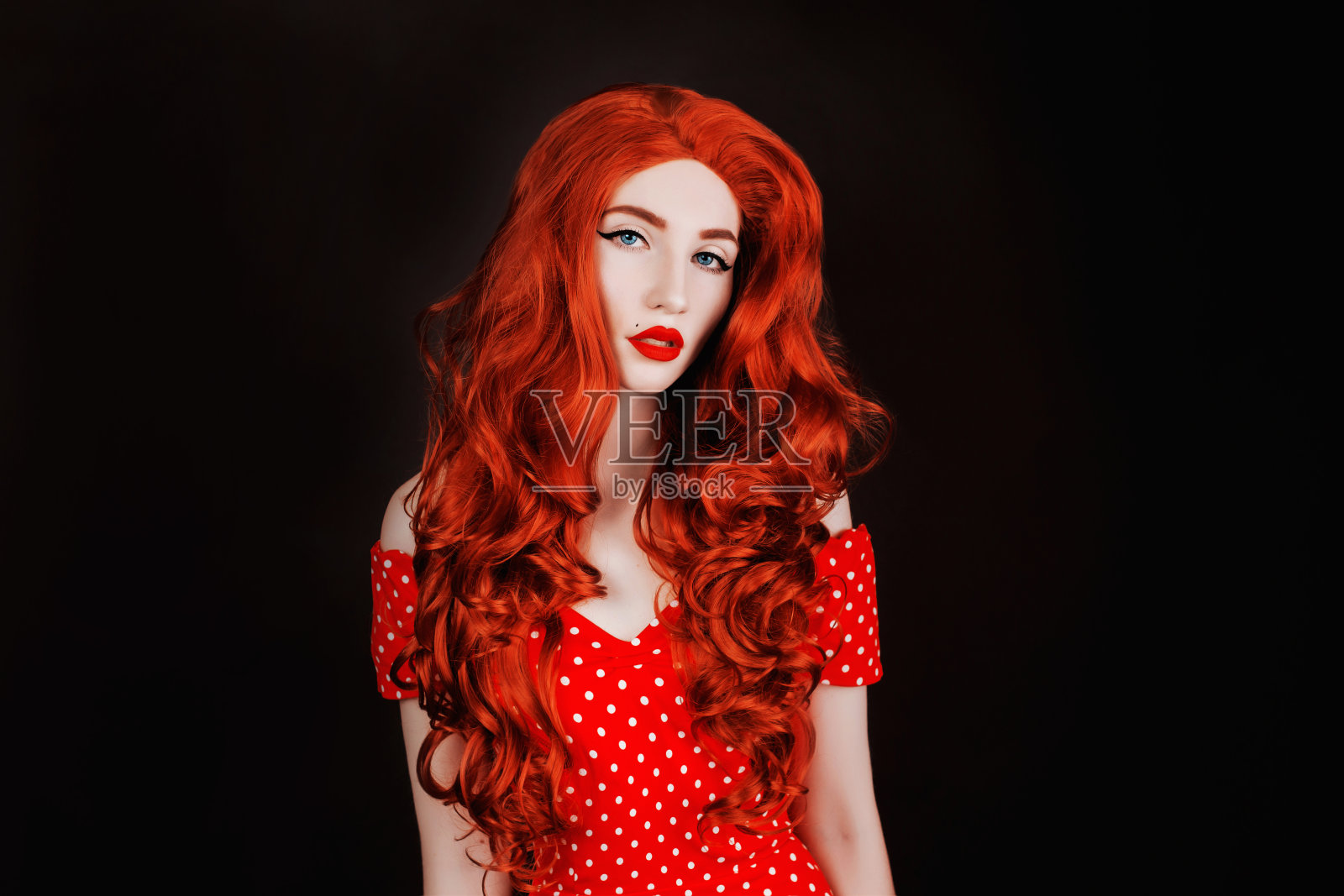 黑背景上的红发女子,皮肤苍白,嘴唇红润。穿复古裙的铜色头发女孩。红色卷曲的假发。红色头发的女人。长的卷曲的假发。大量的卷发。穿着波尔卡圆点裙的复古女士照片摄影图片