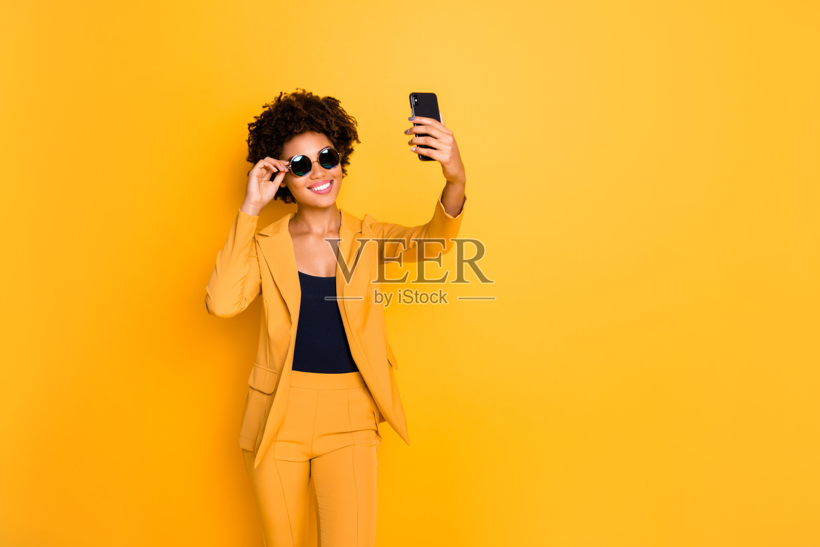 正面开朗的深色皮肤享受假期用她的手机自拍博客时代时尚时尚的服装孤立在黄色背景照片摄影图片