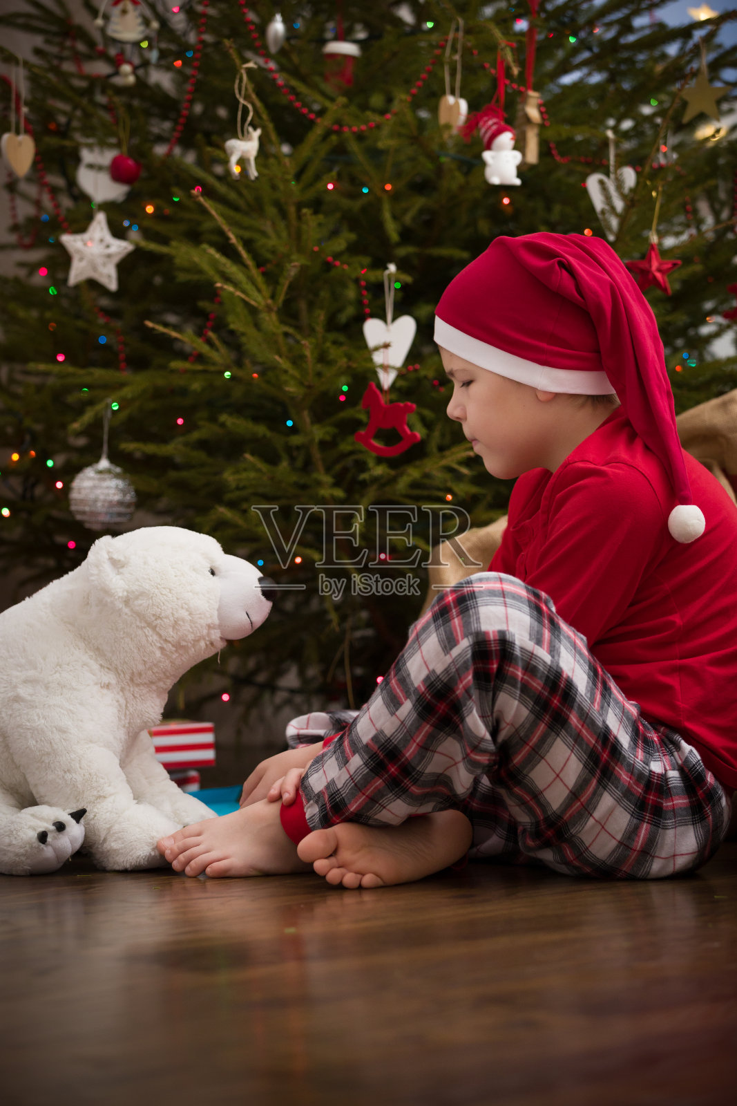 看看可爱的小男孩和他的玩具熊坐在圣诞树旁享受庆祝。假期。新年快乐。照片摄影图片