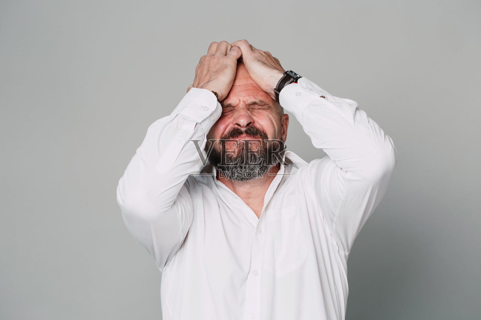 一名英俊严肃的中年男子,穿着一件白色衬衫,很生气,在灰色的背景上,他的手靠近自己的脸照片摄影图片