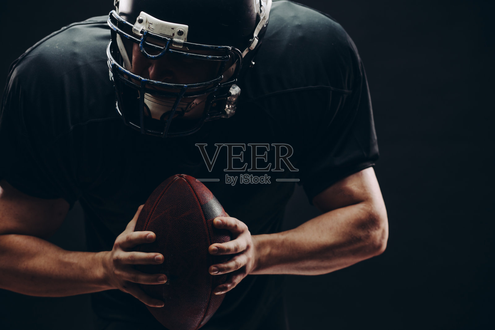 戴头盔和盔甲的美国足球运动员,用球对着黑墙照片摄影图片