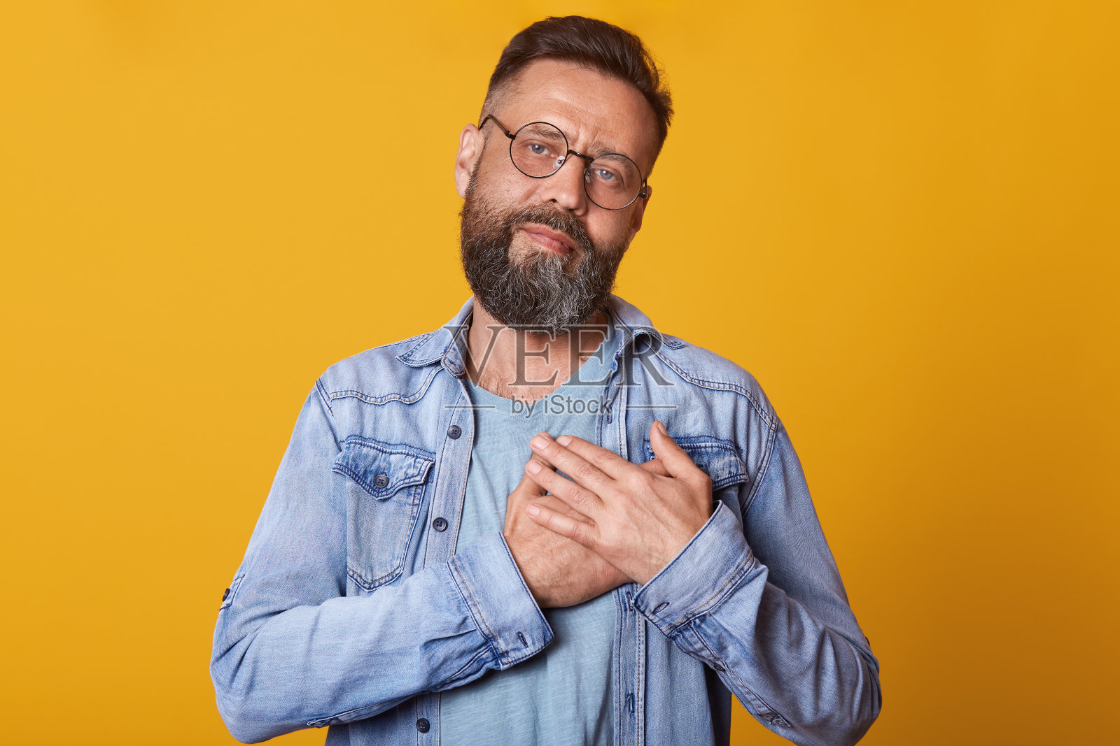 图片中的大胡子男子戴着圆形光学眼镜,双手放在胸前,穿着时尚的牛仔夹克,在黄色的工作室背景下摆出孤立的姿势,表达了他的爱和感激。照片摄影图片