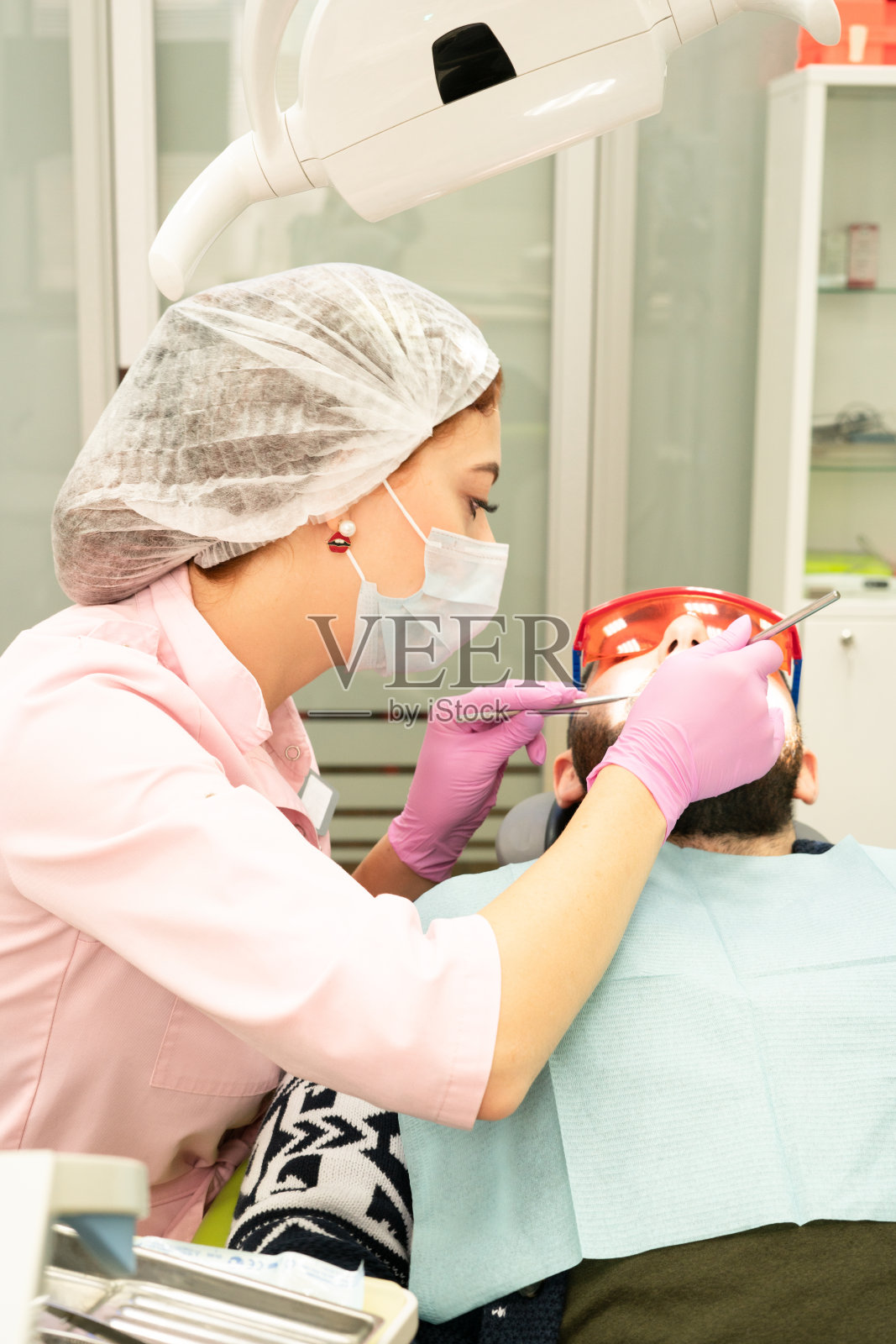 牙医一个年轻的女人给一个男人看病。医生使用一次性手套、口罩和帽子。牙医在病人嘴里工作,使用专业工具照片摄影图片