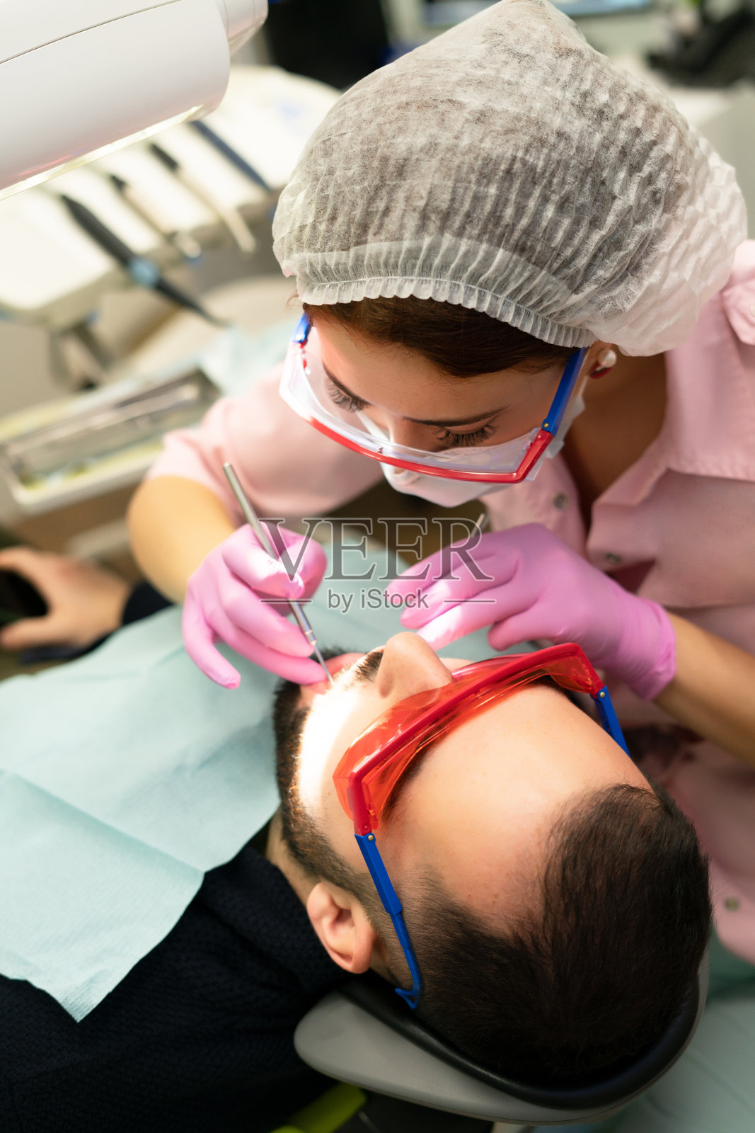 牙医一个年轻的女人给一个男人看病。医生使用一次性手套、口罩和帽子。牙医在病人嘴里工作,使用专业工具照片摄影图片