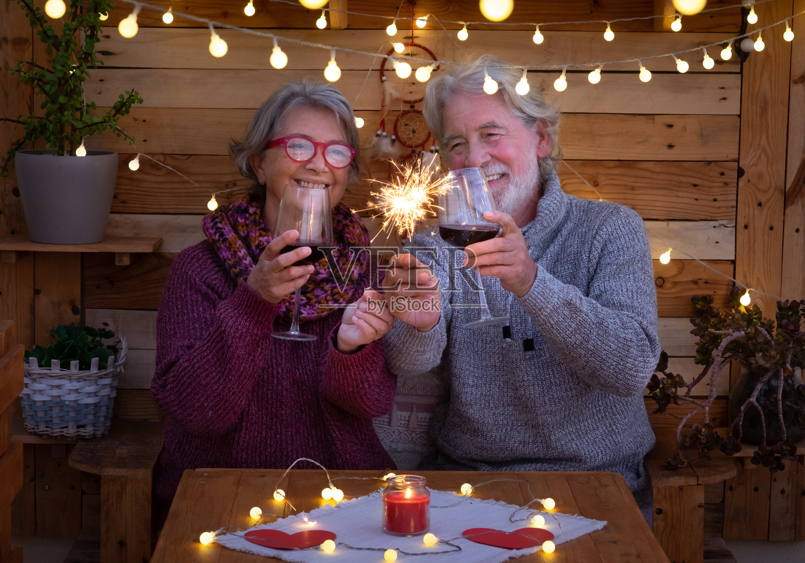一对面带微笑、闪闪发光的老年夫妇坐在户外一个浪漫的木制角落里。黄色的灯光。两杯红酒来庆祝一个积极的时刻照片摄影图片