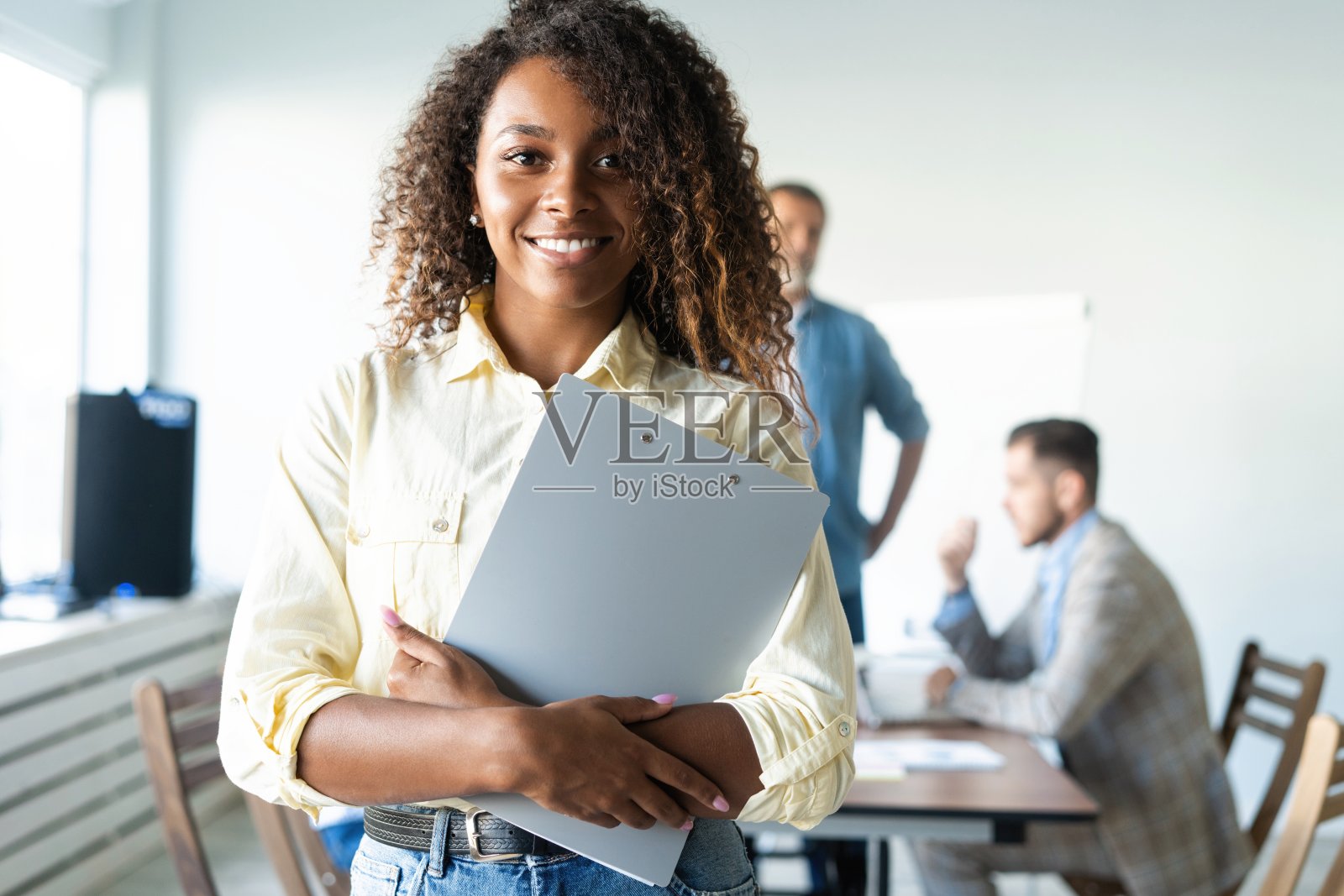 美丽年轻的非洲职业女性站在办公室里,而工人们在后台开会。照片摄影图片