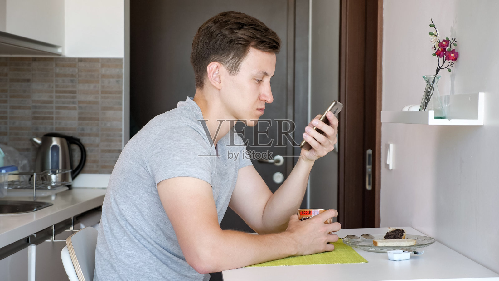 一名男子坐在一间小厨房里,一边喝茶,一边浏览手机。照片摄影图片