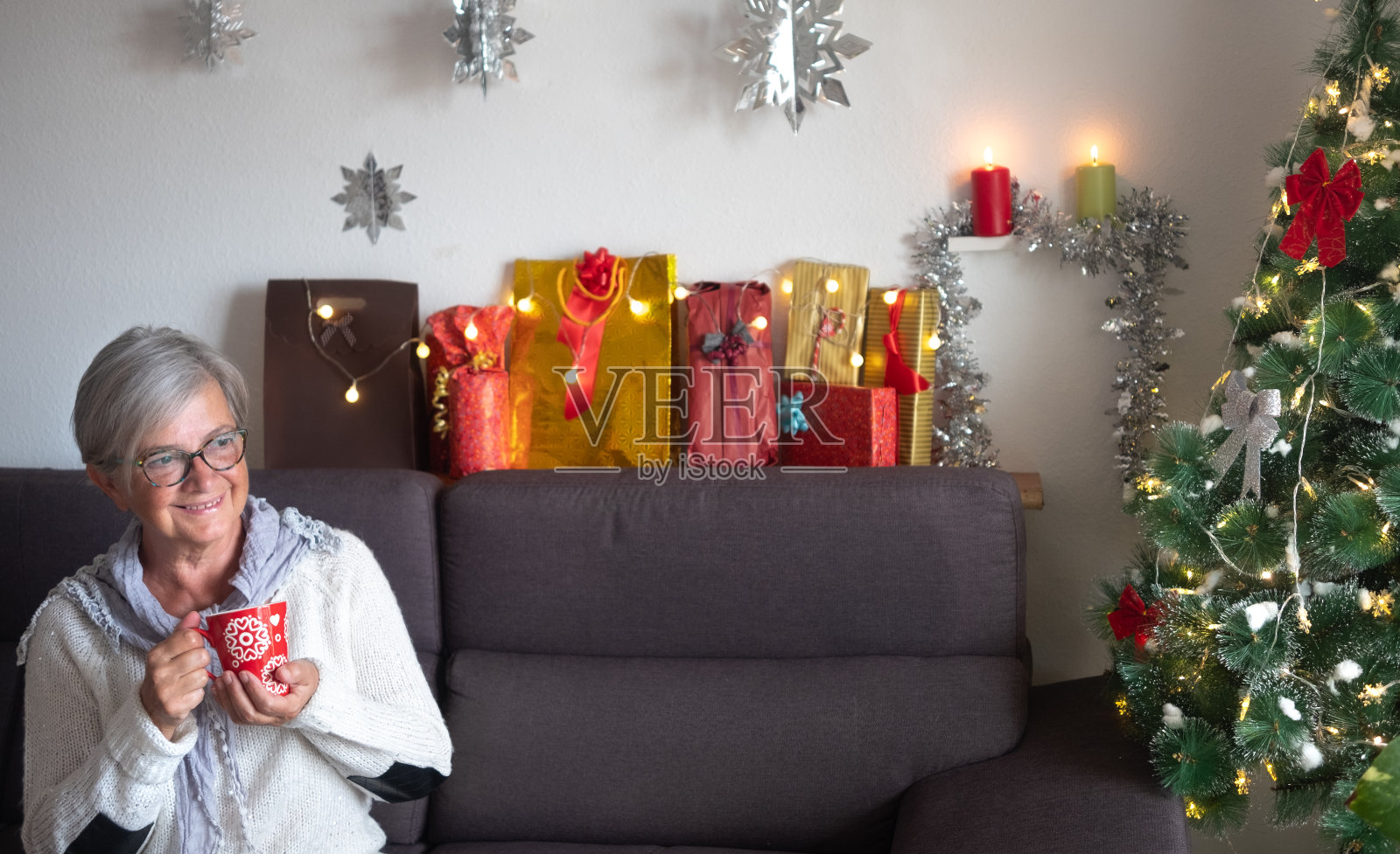 前面的观点,一个年老的女人与灰色头发坐在沙发上的圣诞树和礼物的背景。一位老人喝着茶放松照片摄影图片