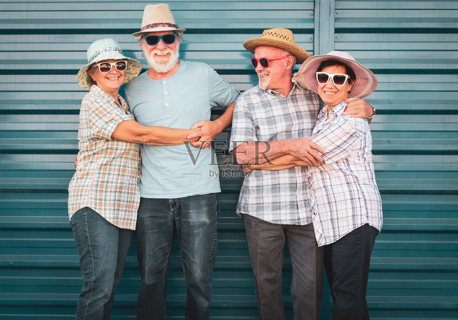 四名老人面带微笑,站在一堵蓝色的墙上互相拥抱。有胡子的男人。所有人都戴着帽子。退休人员的笑脸是欢快和轻松的。照片摄影图片