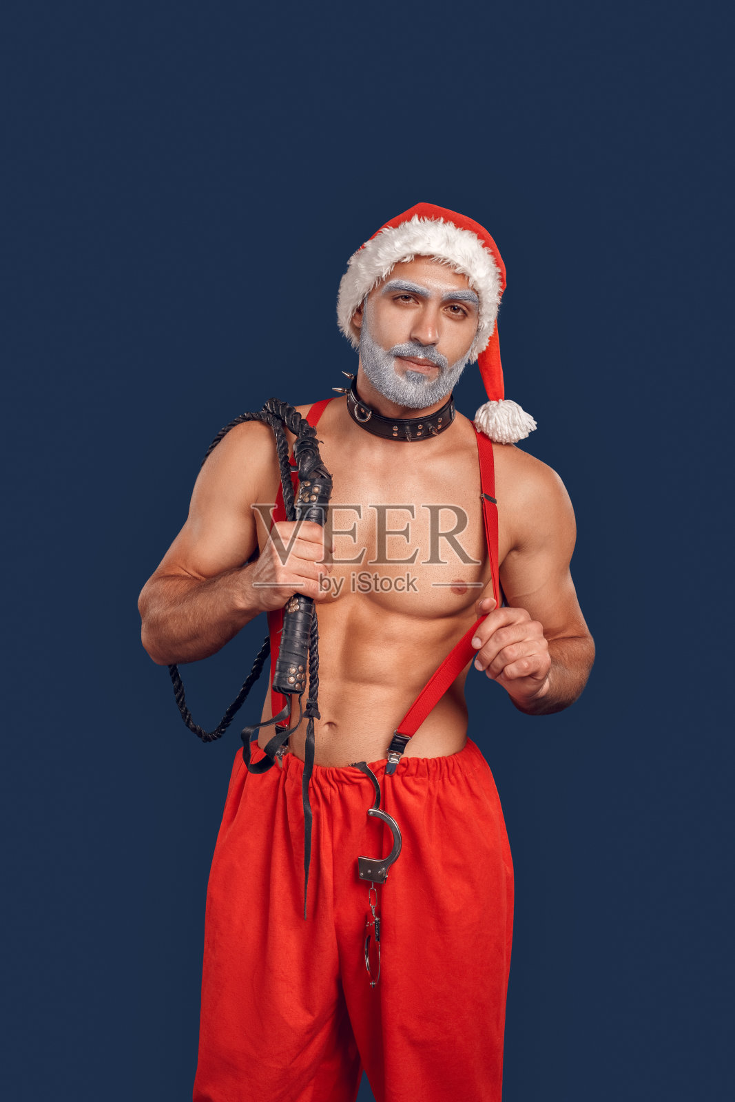 圣诞节自由泳。年轻的圣诞老人裸露肌肉上半身在帽子和狗项圈站在孤立的蓝色持有手铐和鞭子摆姿势微笑自信照片摄影图片
