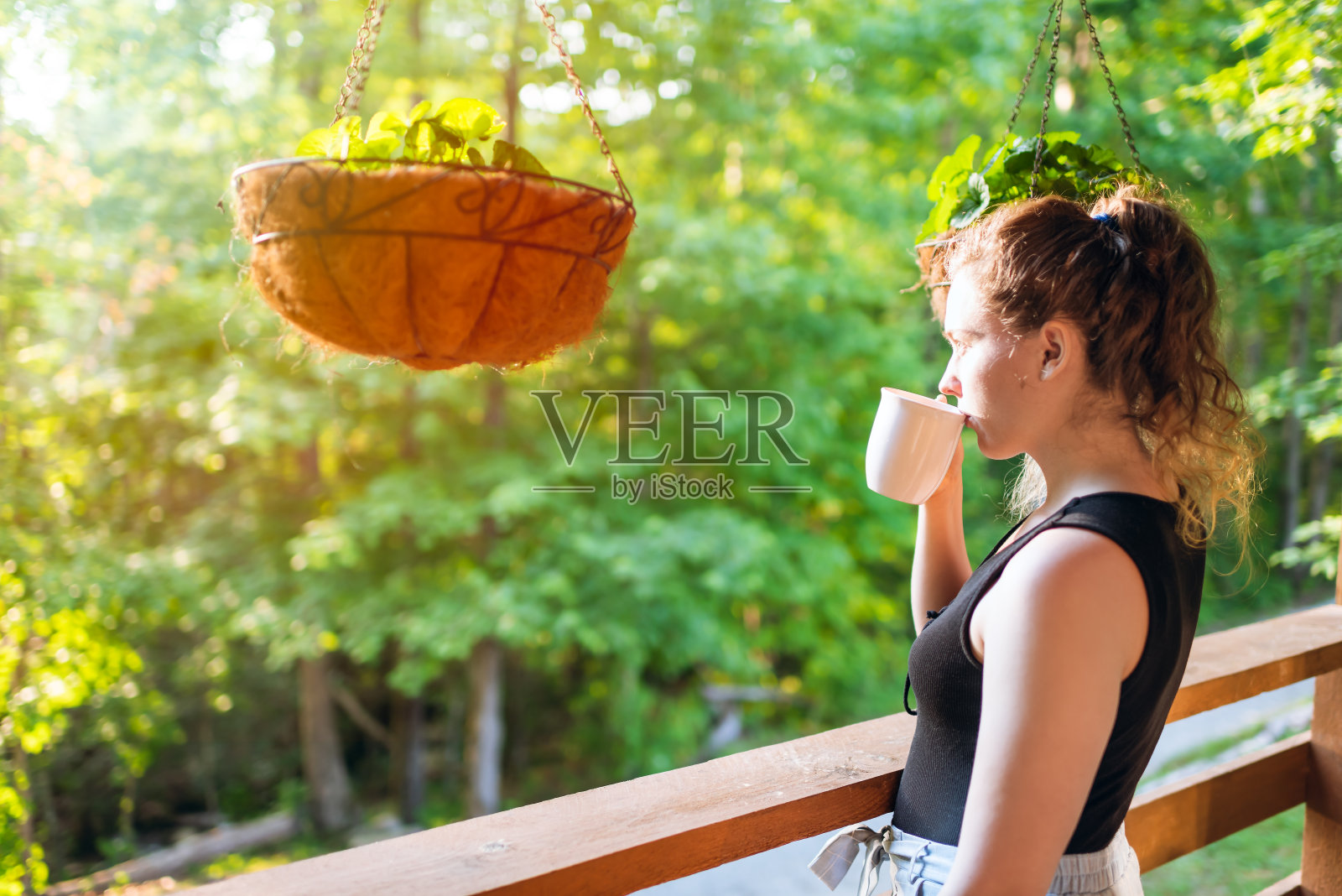 夏季悬挂盆栽,女人站在房屋门廊上,在早晨的木屋小屋里喝茶或咖啡照片摄影图片