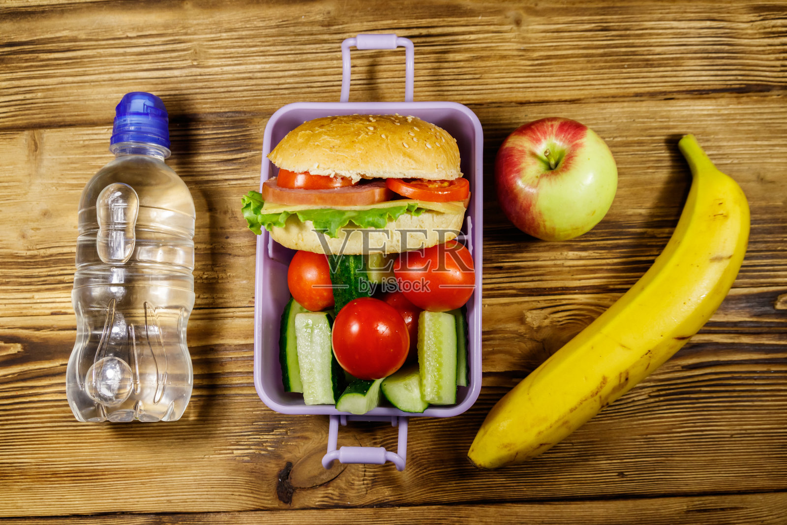 木桌上放着一瓶水、苹果、香蕉和一盒汉堡和新鲜蔬菜。俯视图照片摄影图片
