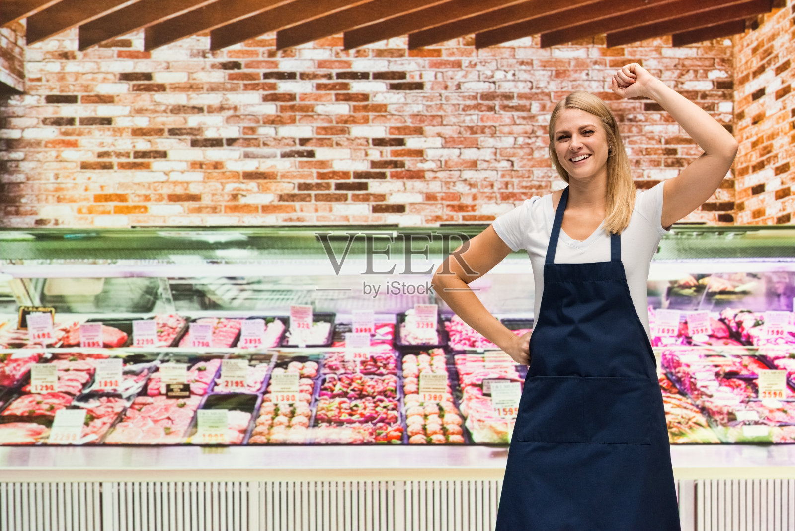 一个人/腰部以上20-29岁的成年人漂亮的金发/长发白人女性/年轻女性屠夫/店主站在肉店/熟食店/超市/市场-零售空间/商店的冷藏区照片摄影图片