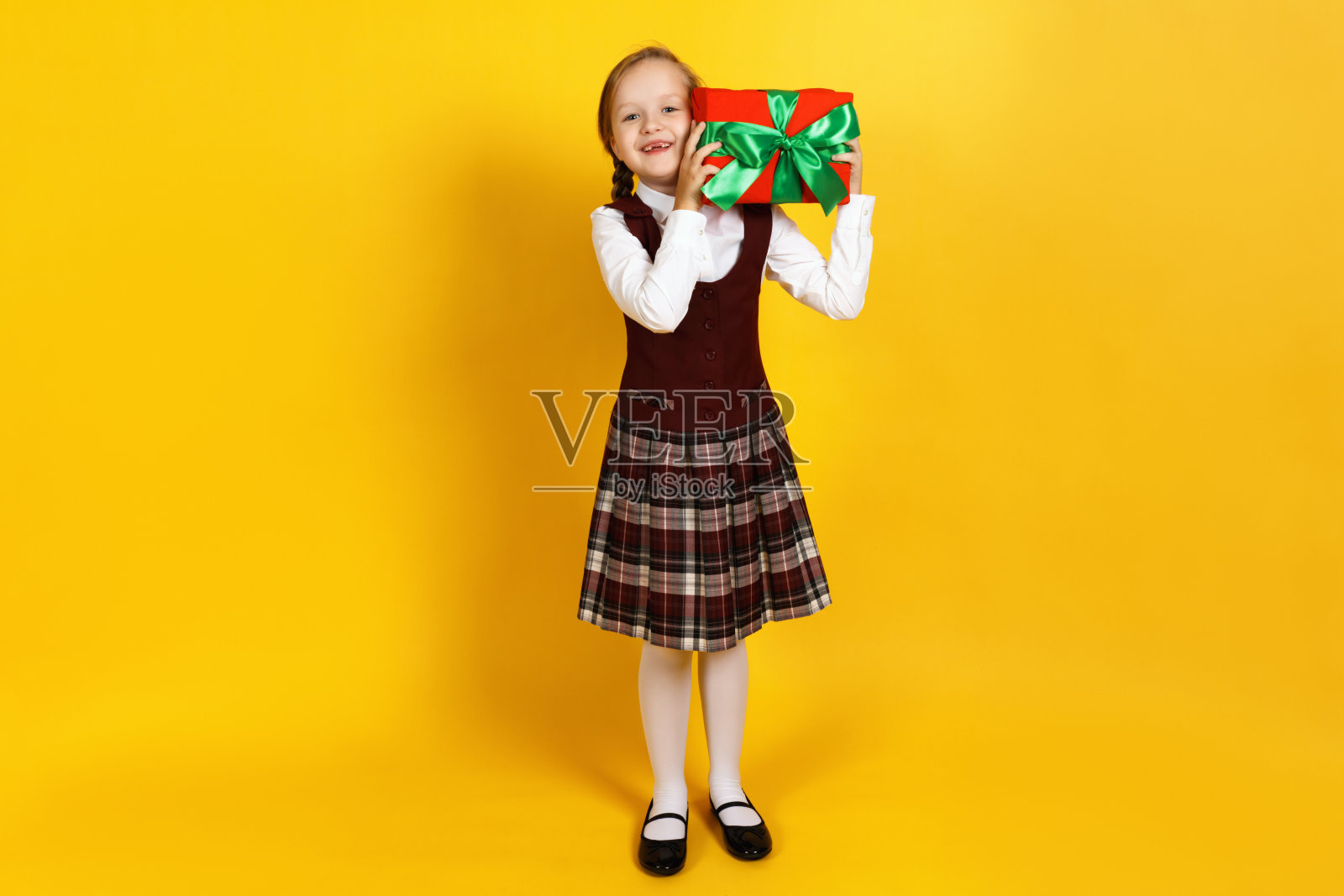 快乐快乐的小女孩拿着一个装着礼物的盒子。一个梳着辫子的孩子,穿着白色衬衫,黄色背景上穿着紫红色的背心裙照片摄影图片