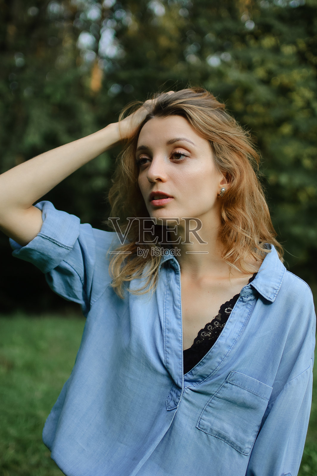 性感的金发女孩穿着蓝色休闲牛仔裤衬衫和黑色内衣摆姿势在绿色的树木背景的公园照片摄影图片