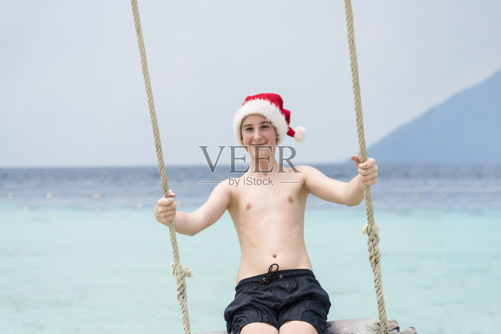 戴着圣诞老人帽子的小男孩在海上荡着绳索照片摄影图片