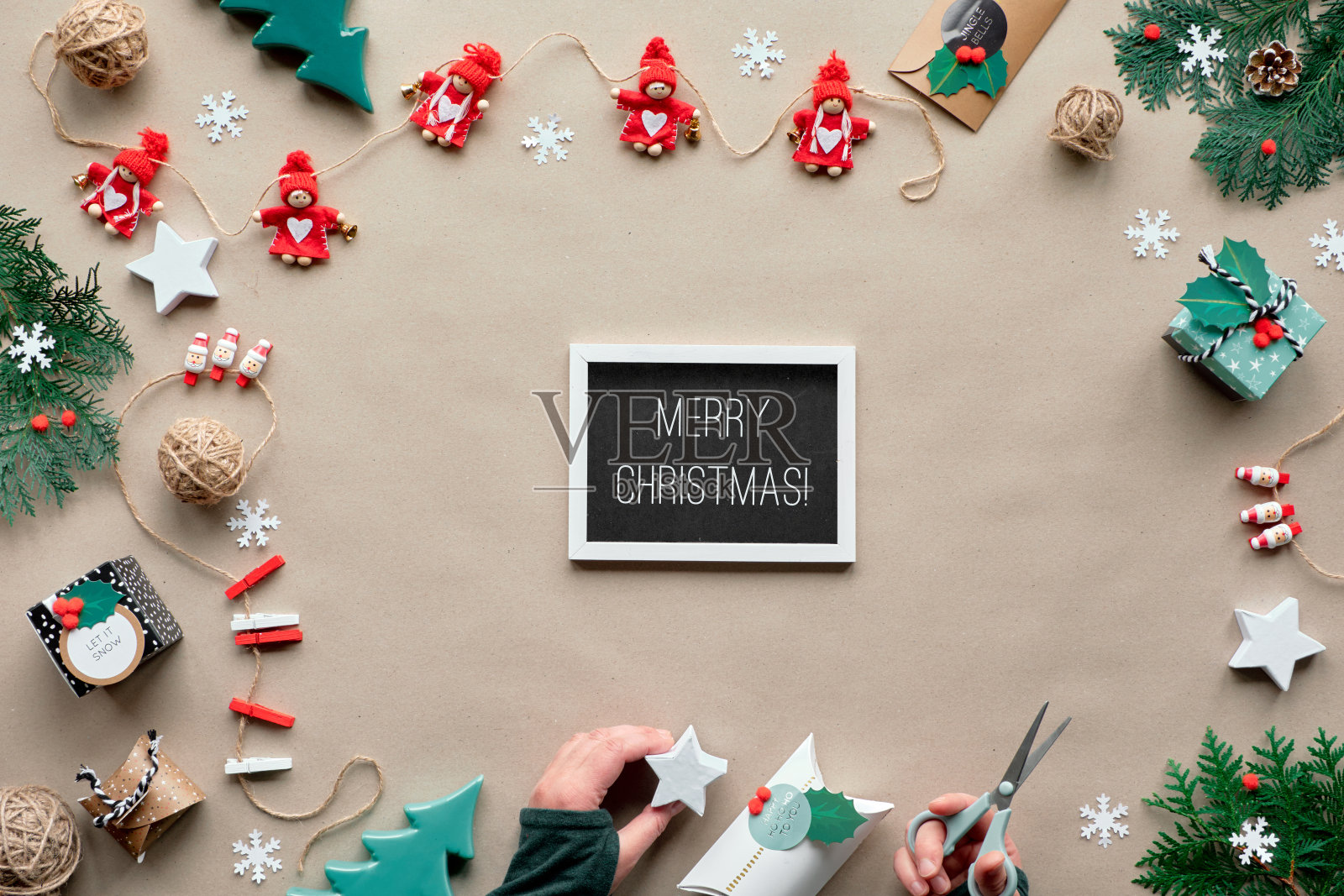 创意手工装饰,零浪费圣诞框架。平铺,牛皮纸俯视图。纺织品小饰品,手里拿着装饰好的礼盒。生态友好的圣诞节。在黑板上写上“圣诞快乐”。照片摄影图片