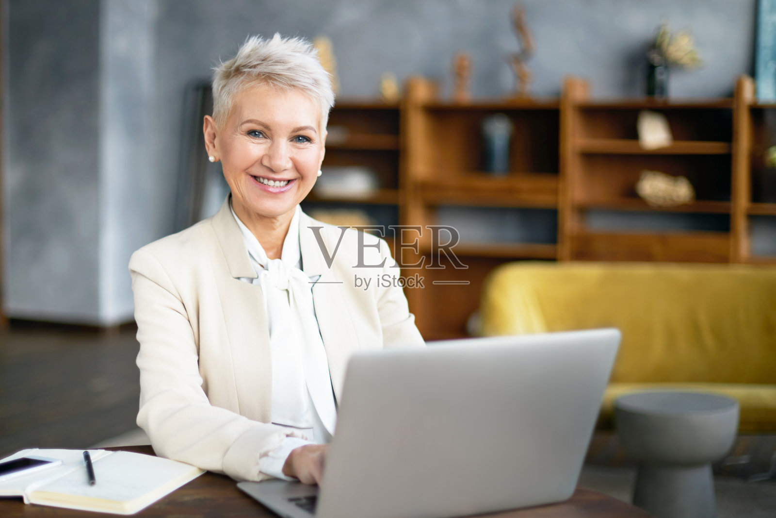 50岁的成功女商人,一头金色短发,容光焕发的笑容,用笔记本电脑上网交流,坐在办公桌前工作,输入商业广告。年龄、技术和生活方式照片摄影图片