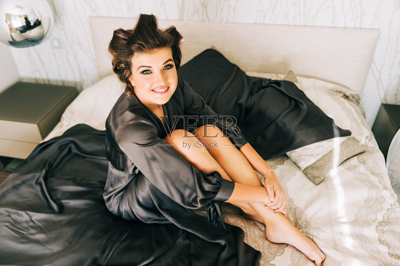 室内肖像的快乐年轻女子放松在卧室,穿着灰色睡衣,头上卷发器照片摄影图片