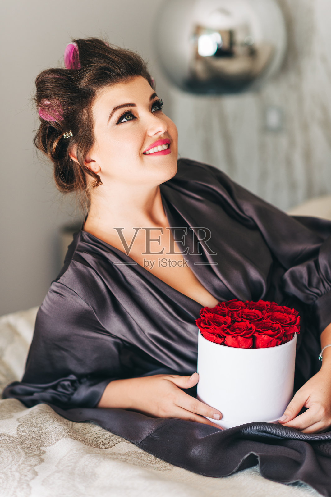 室内肖像的快乐年轻女子放松在卧室,穿着灰色的睡衣,头上的卷发器,拿着礼物盒与红色的稳定玫瑰照片摄影图片