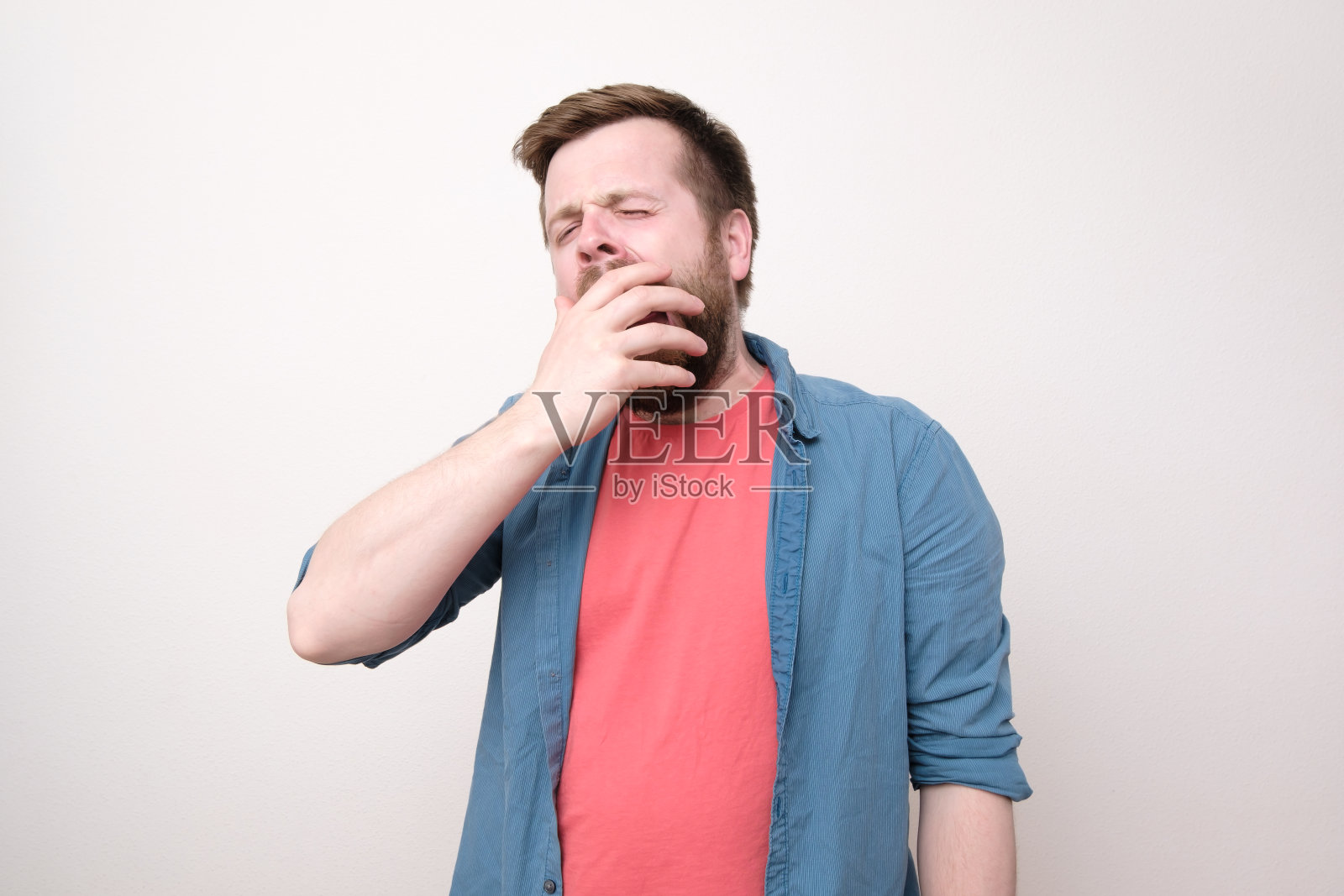 一个留着胡子的欧洲年轻人昏昏欲睡,打了个呵欠,用手捂着嘴。孤立在白色背景上。照片摄影图片