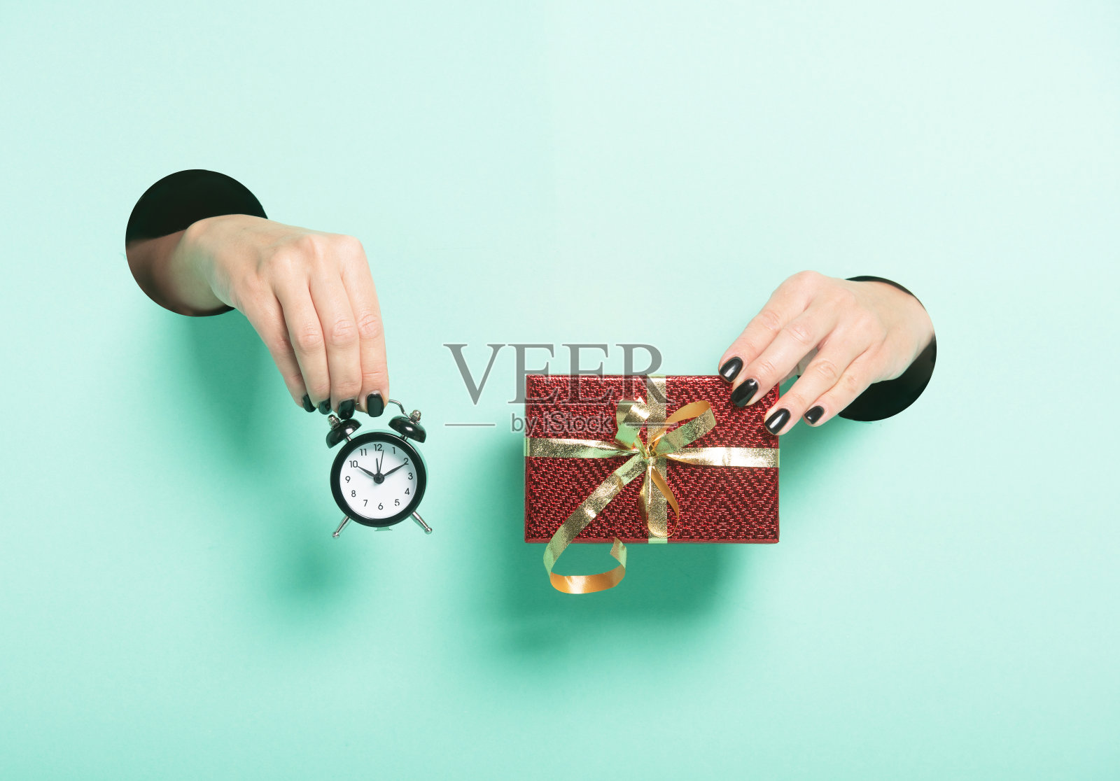 女人的手拿着红色的礼物和闹钟通过一个洞的霓虹灯薄荷背景。极简创意孤立的销售理念。照片摄影图片