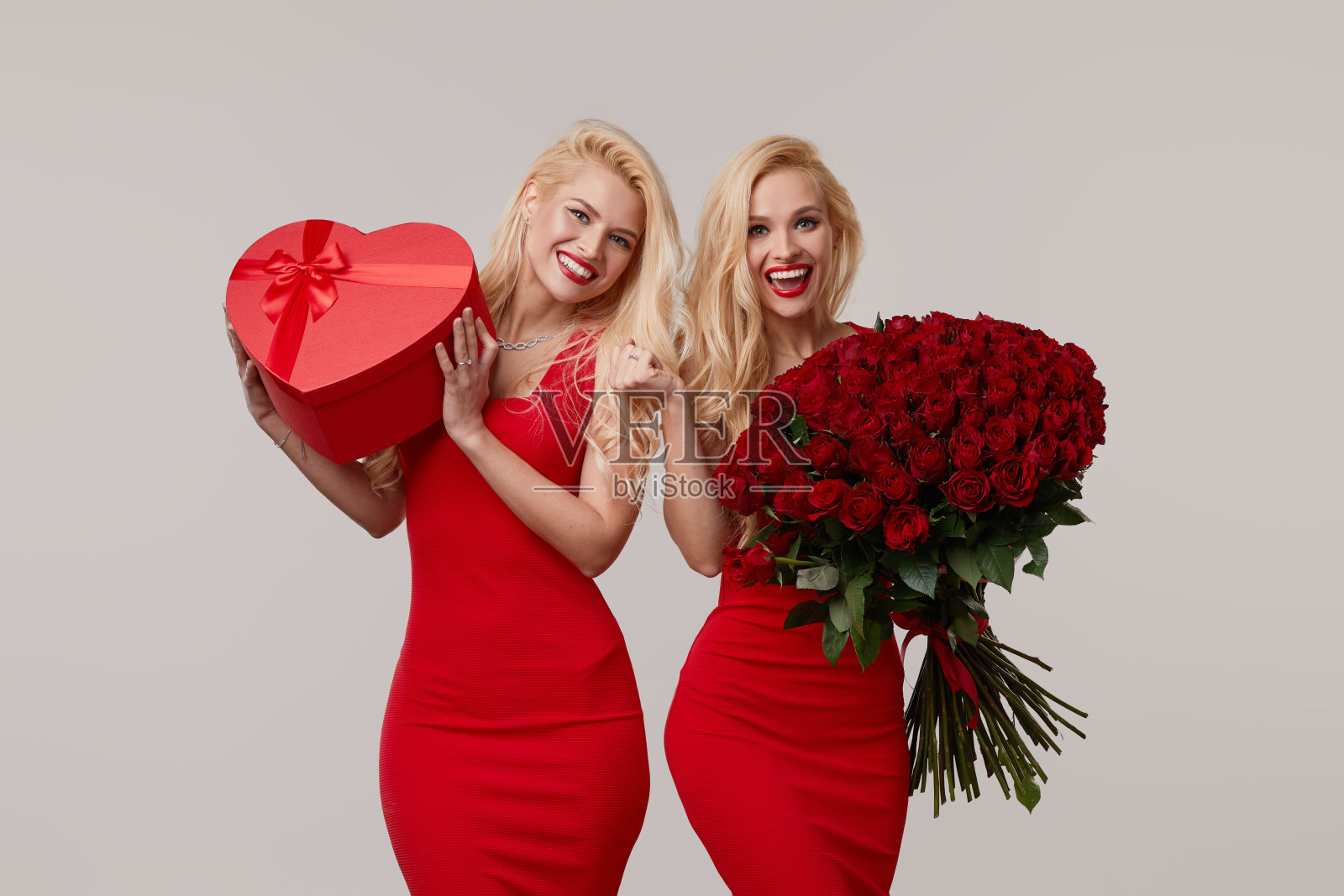 两个快乐的年轻双胞胎女人拿着一大束红玫瑰和一个红色的心型盒子。这个女孩在情人节或3月8日被求婚,并被赠送了一枚结婚戒指照片摄影图片