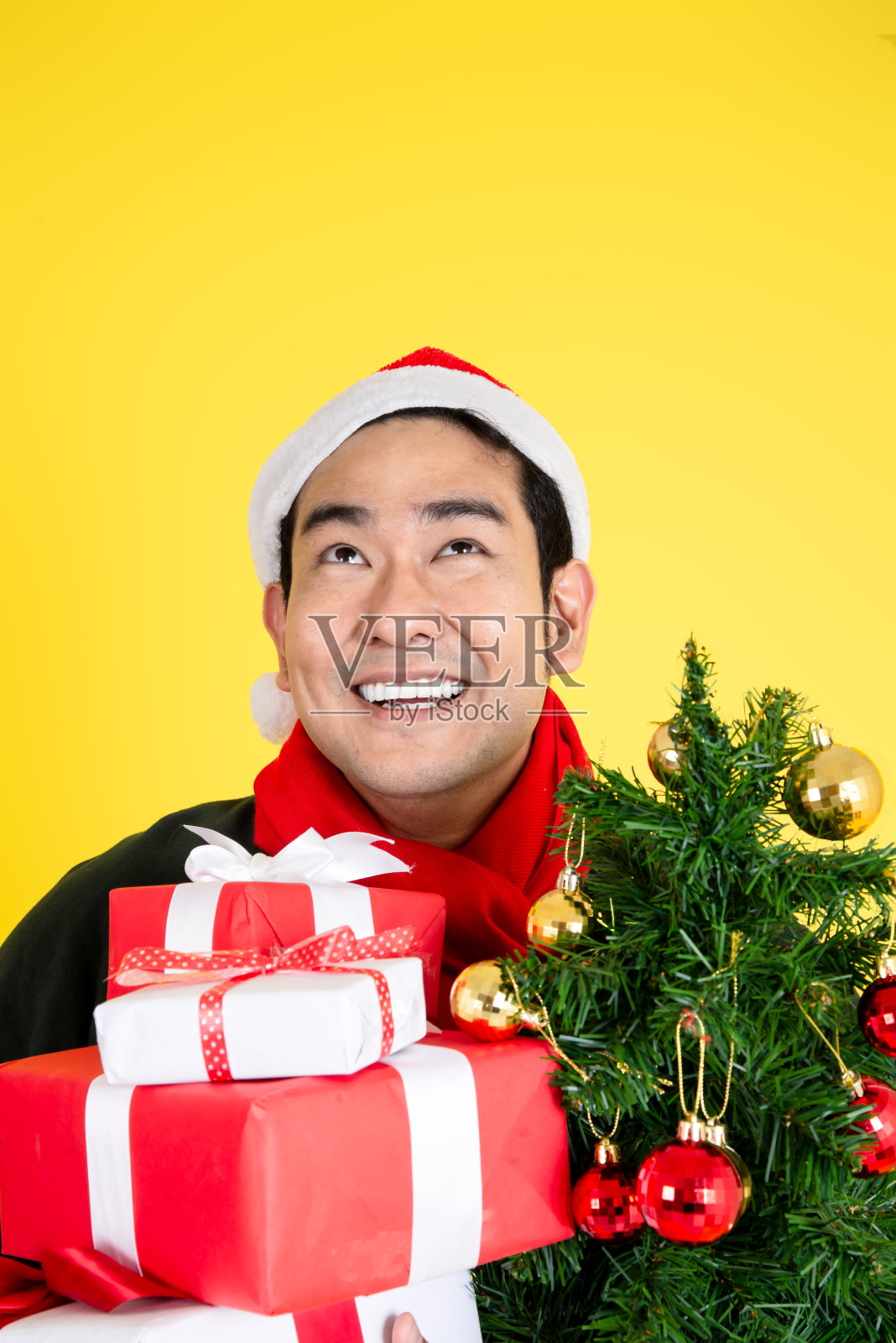 快乐的亚洲男人戴着圣诞帽,拿着礼品盒,在黄色背景上向上看照片摄影图片