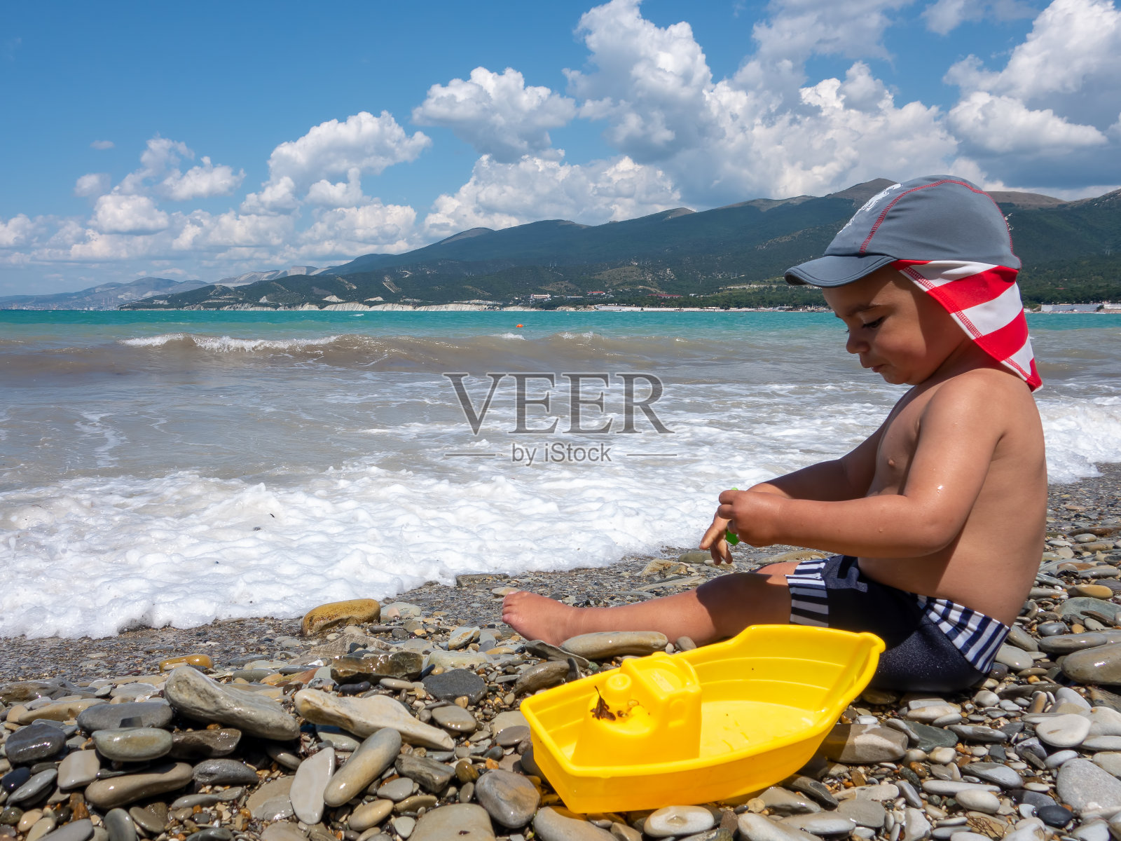 戴着巴拿马帽的小男孩在海滩上用黄色玩具船和绿色肩胛骨在海边玩耍照片摄影图片