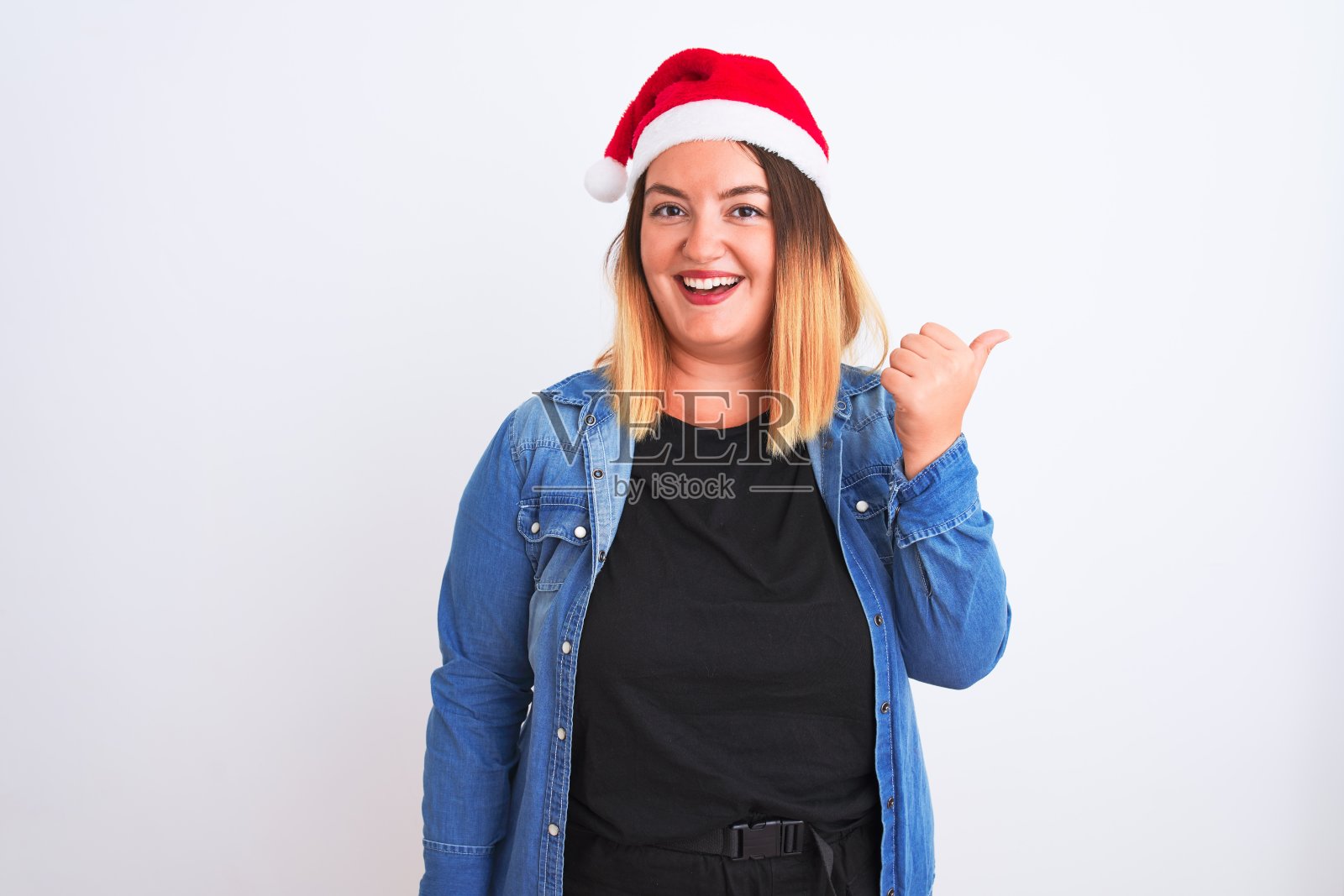 年轻美丽的女人戴着圣诞老人的帽子站在孤立的白色背景微笑与快乐的脸看和指着一边拇指向上。照片摄影图片