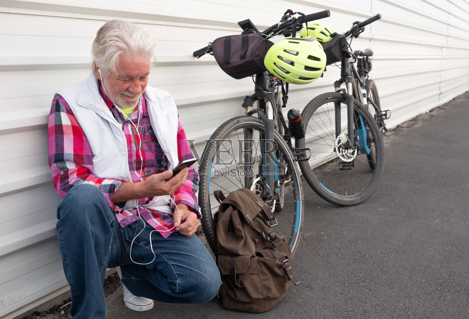 一个戴着耳机的迷人的老人靠在白色的金属墙上。用手机听音乐。他身边的背包。积极的人与他的电动自行车黑色和黄色的头盔照片摄影图片