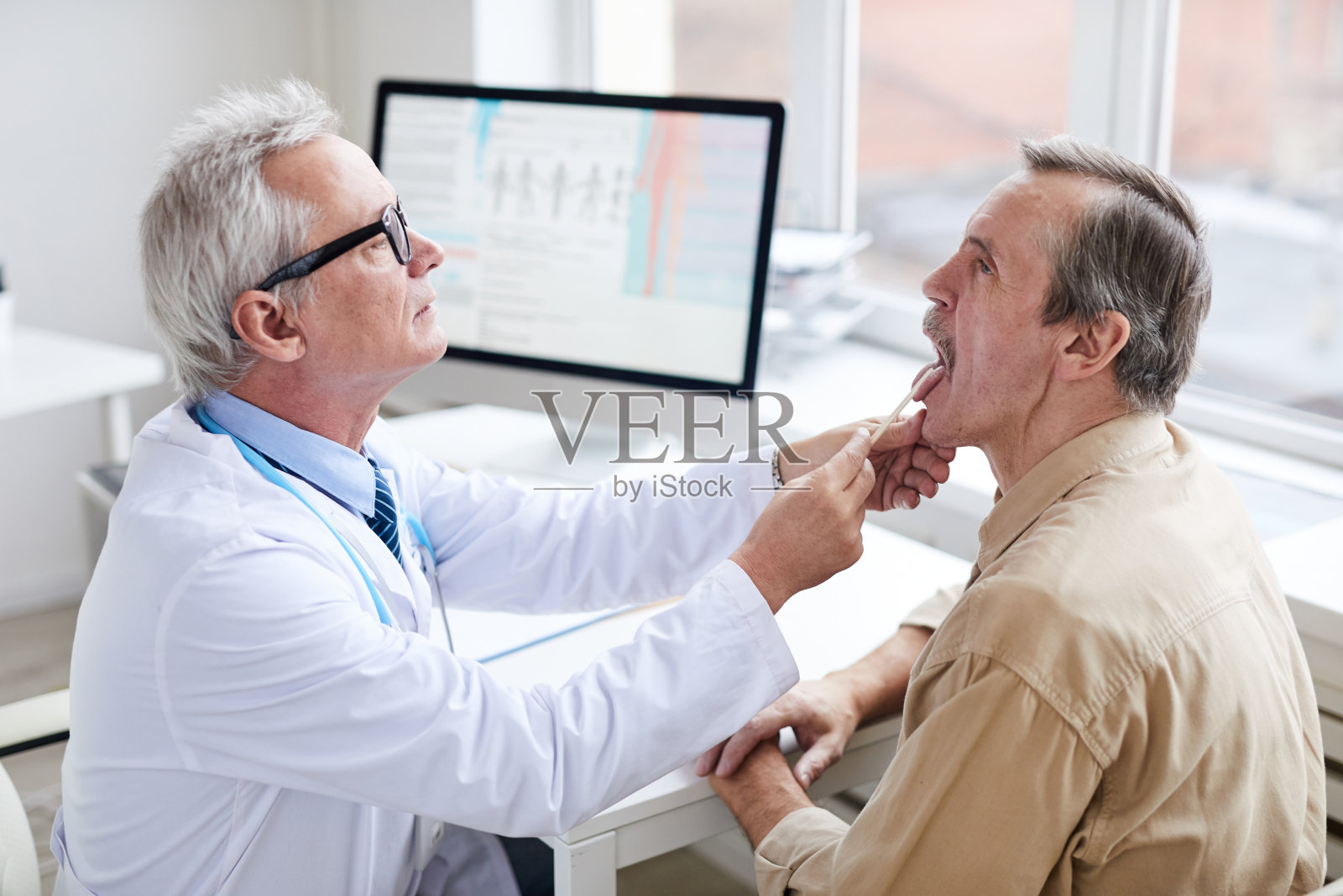 严肃的老年男医生戴着眼镜坐在桌旁,用压舌板给成熟的病人做喉咙检查照片摄影图片