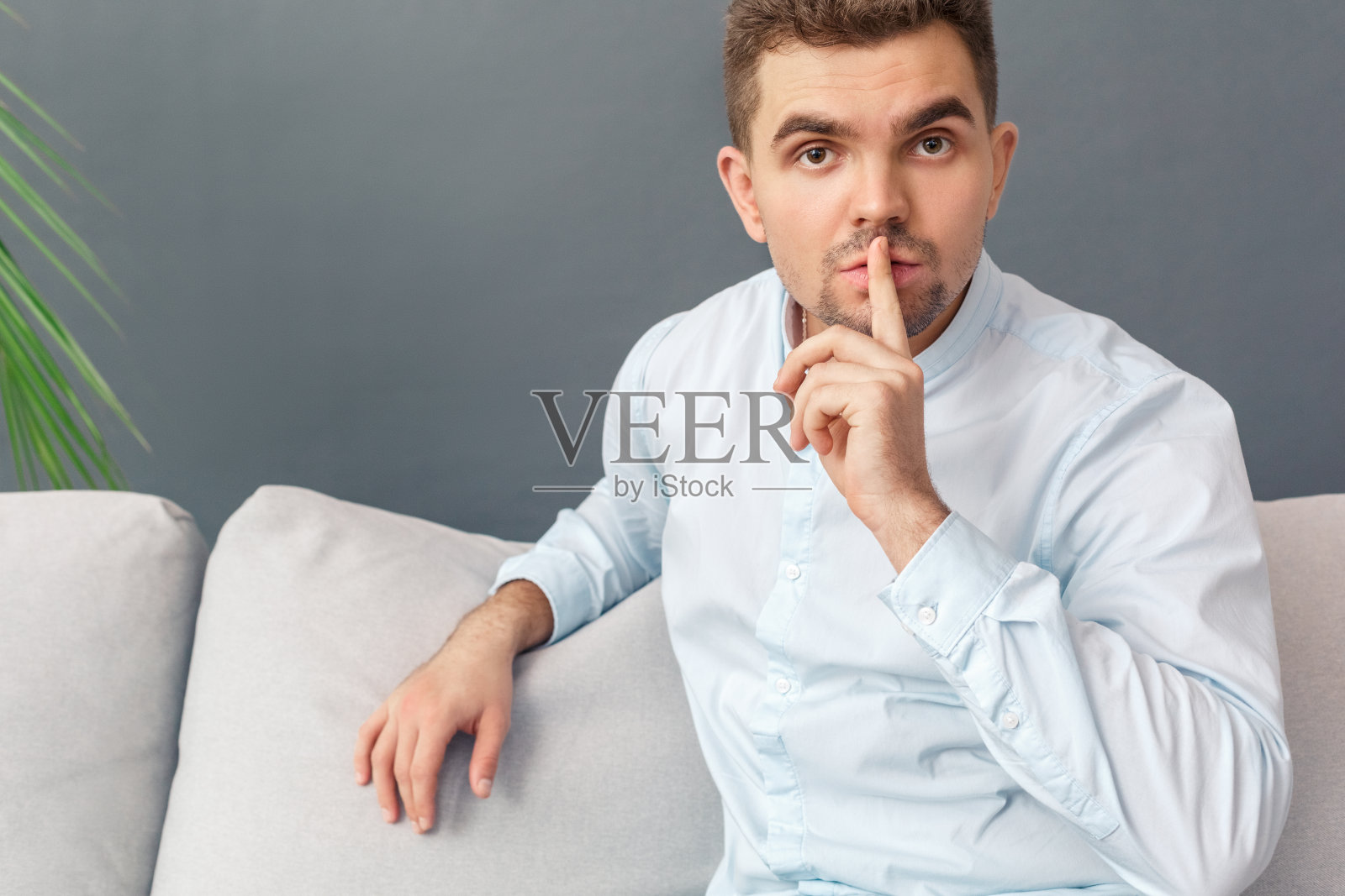 保持秘密。年轻男子坐在沙发上孤立的灰色显示沉默的手势对相机特写照片摄影图片