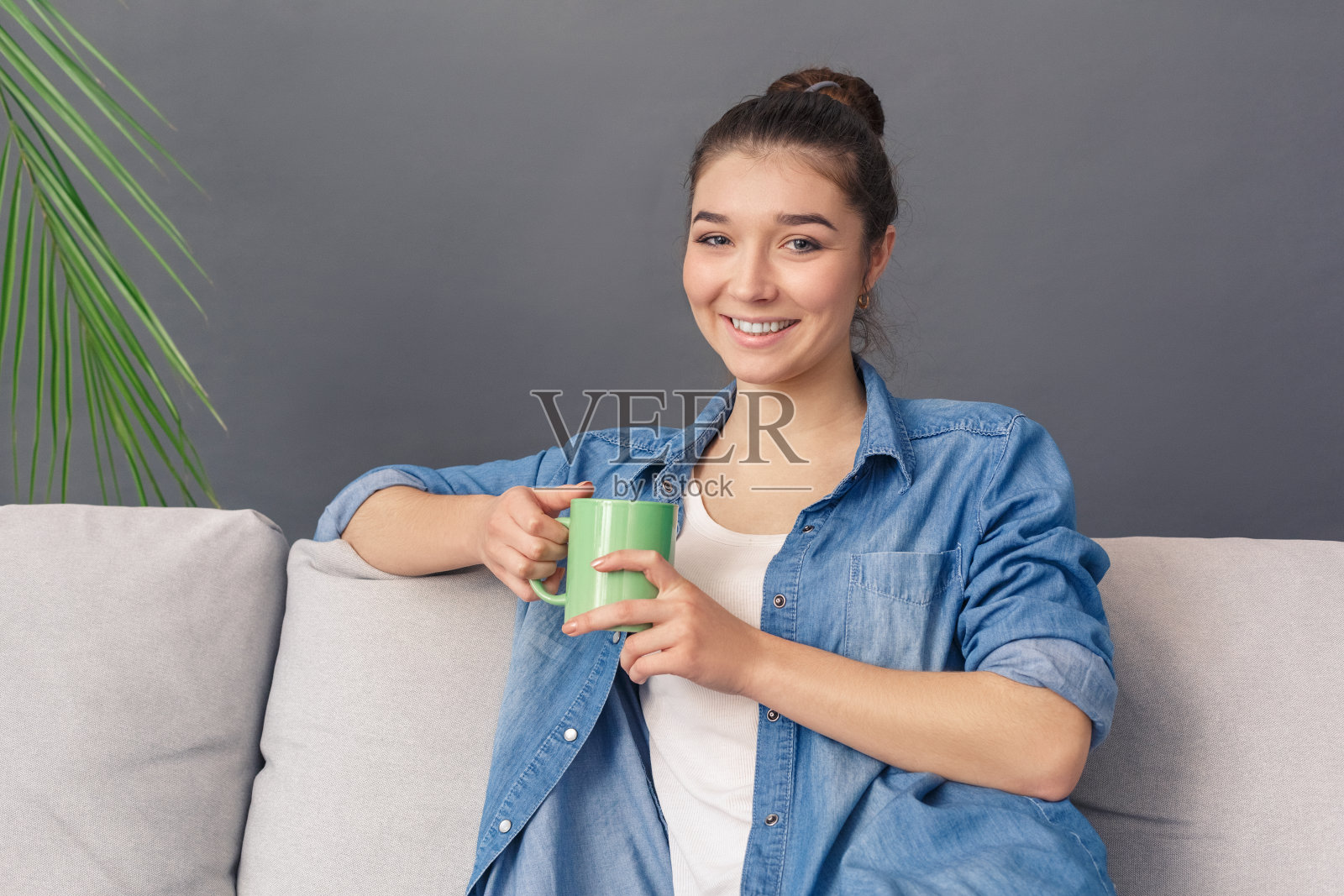 自由泳。年轻女子坐在沙发工作室孤立在灰色看电视喝着热咖啡愉快地对着镜头微笑照片摄影图片