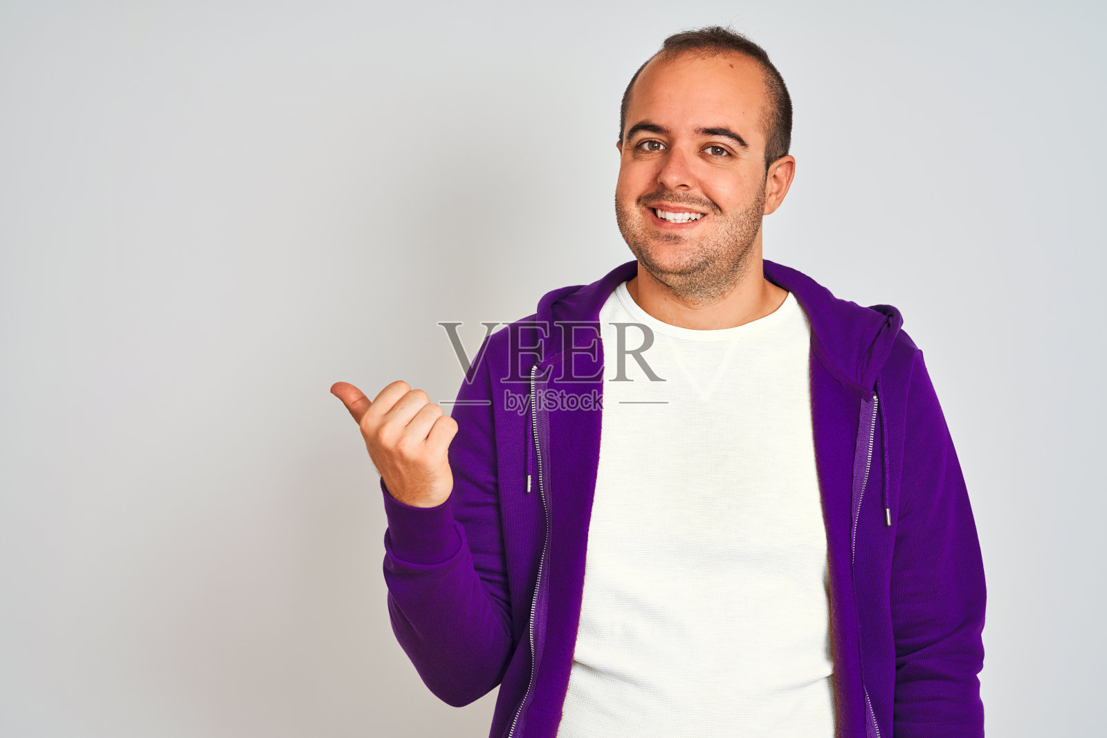年轻男子身穿紫色运动衫站在孤立的白色背景微笑与快乐的脸看和指着一边拇指向上。照片摄影图片